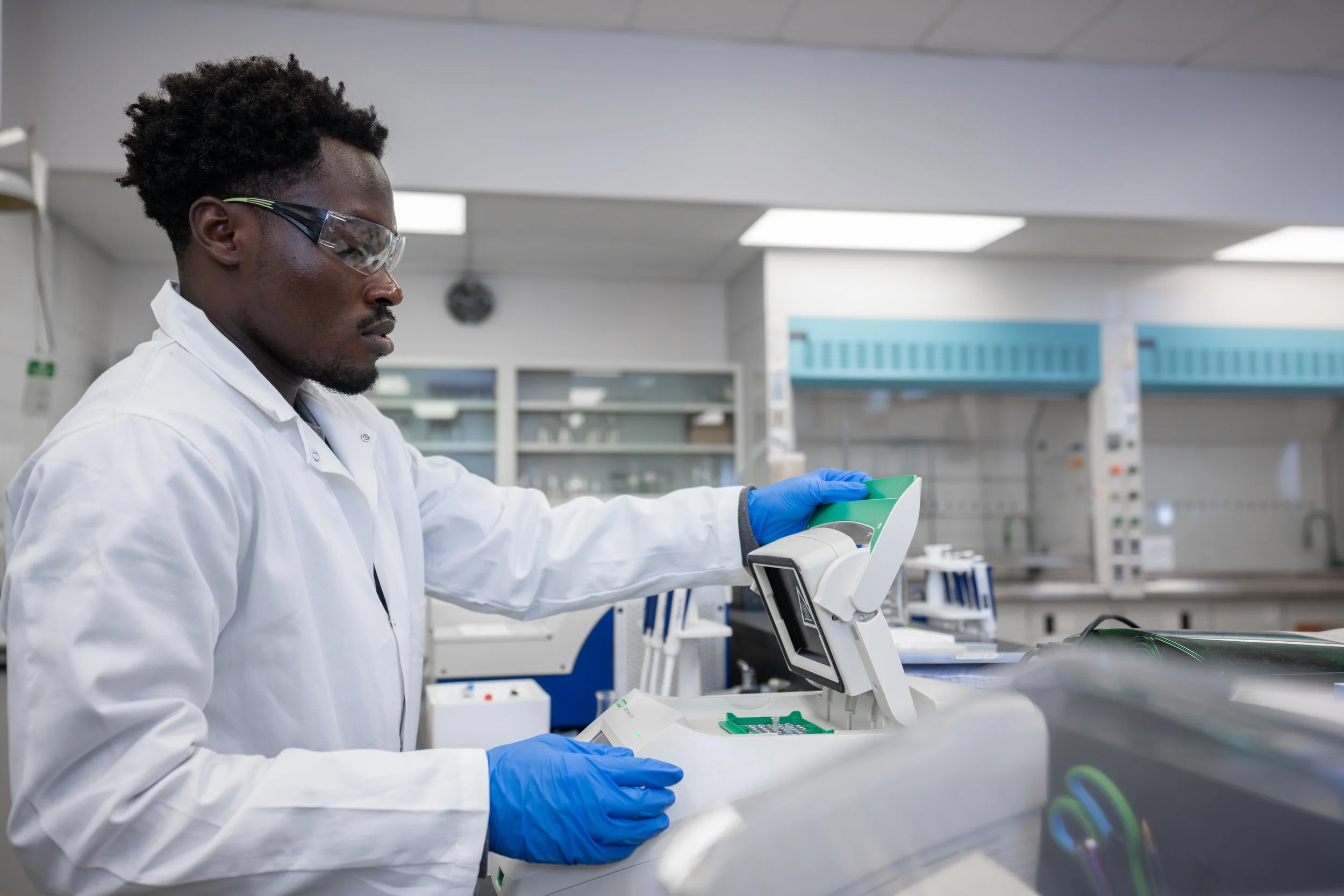 A scientist wearing safety goggles, a white lab coat, and blue gloves working with laboratory equipment in a laboratory.