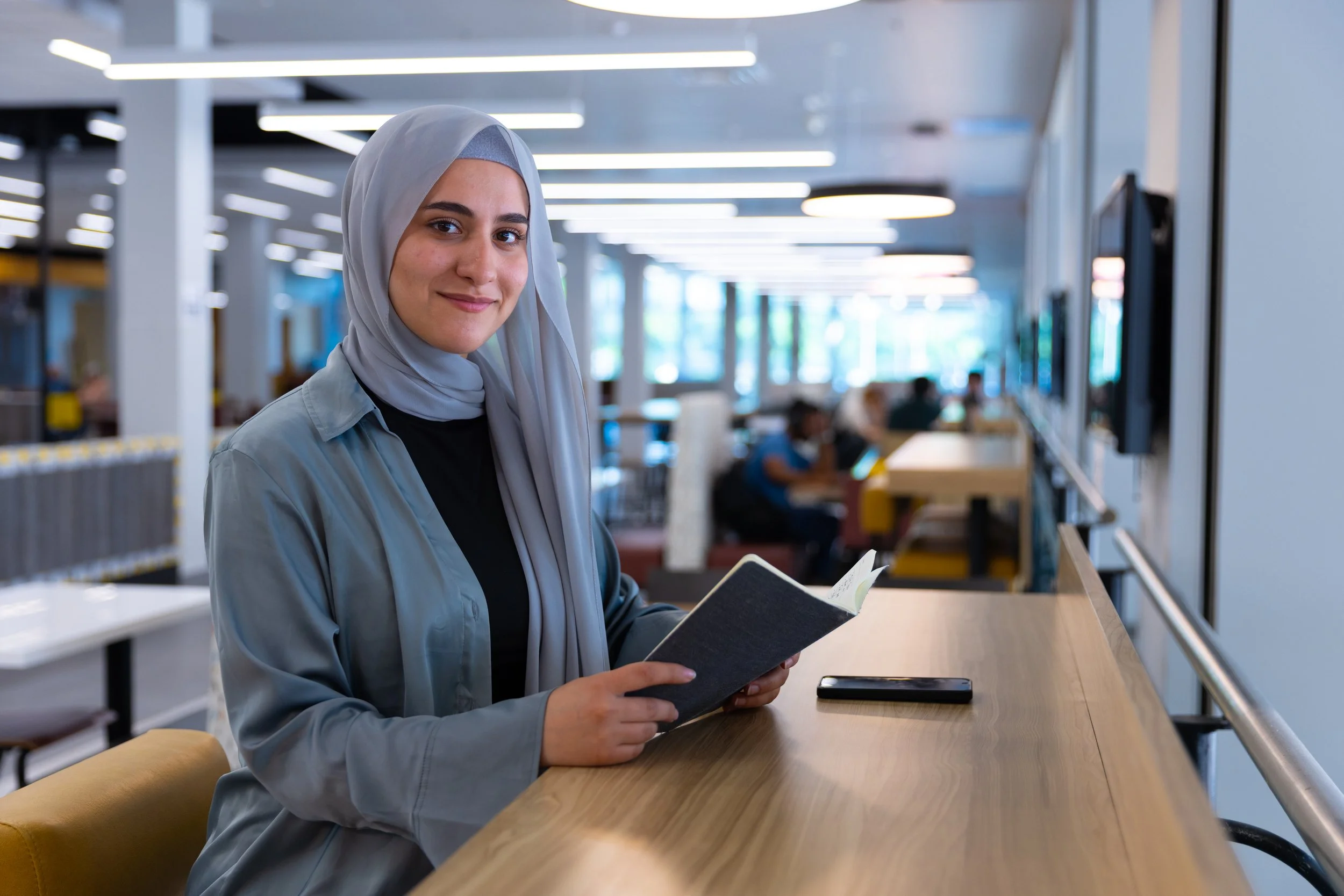 A young woman wearing a gray hijab and gray jacket standing at a wooden counter in a modern, brightly lit library or co-working space, holding an open book and smiling at the camera.