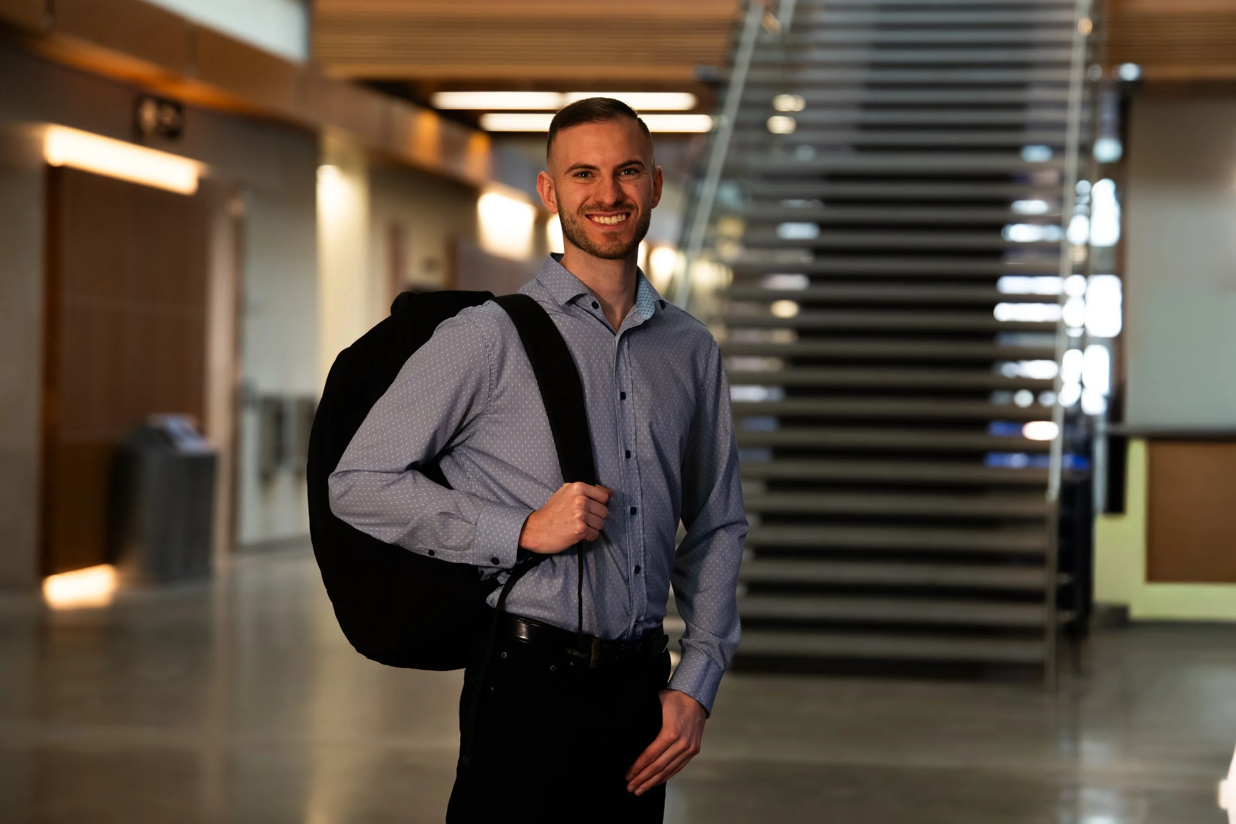 A smiling man in a grey dress shirt with a backpack, standing in a modern indoor setting with stairs in the background.