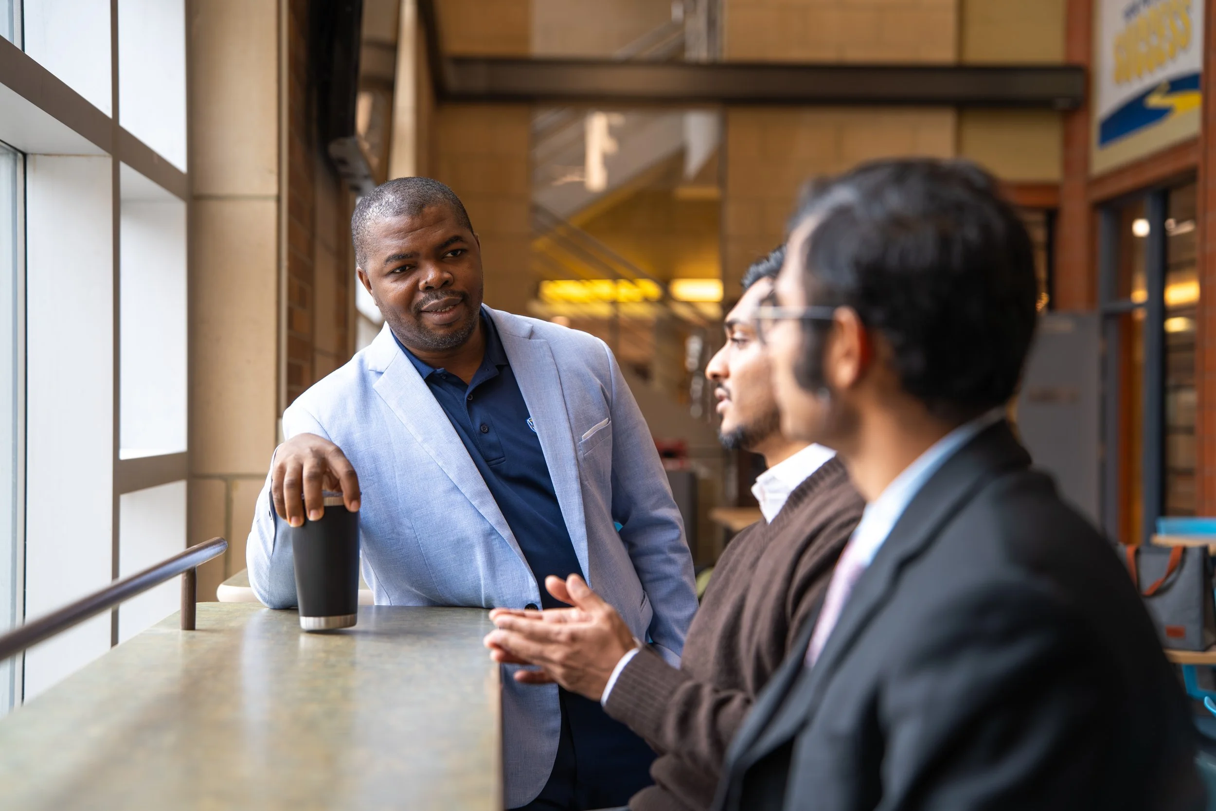 Three men having a conversation at a cafe or restaurant. One man in a light gray blazer is talking, while the other two men in glasses and suits are listening.