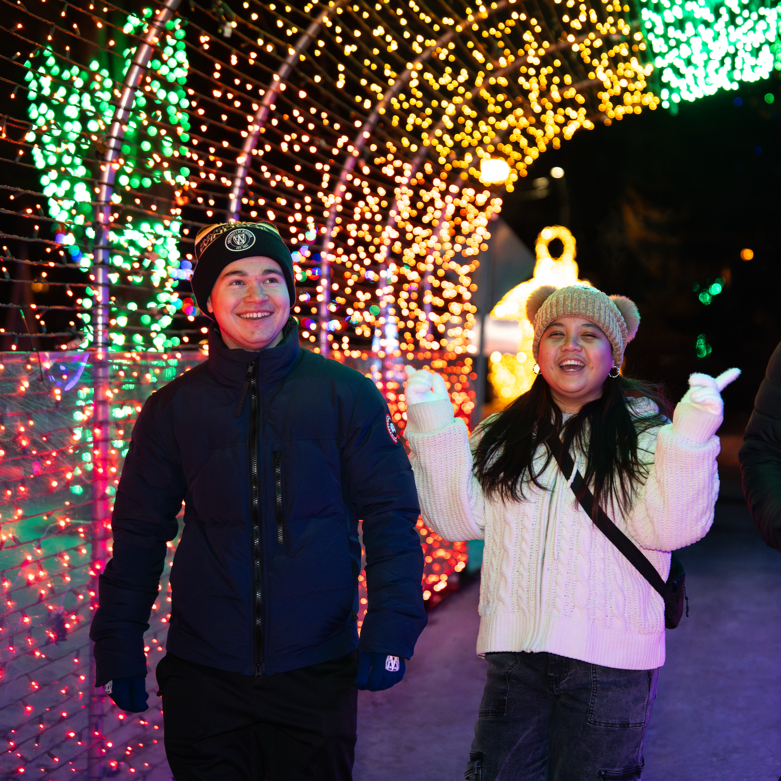 A young man and woman smiling and enjoying a festive outdoor Christmas light display at night. The background features colorful string lights and illuminated decorations.