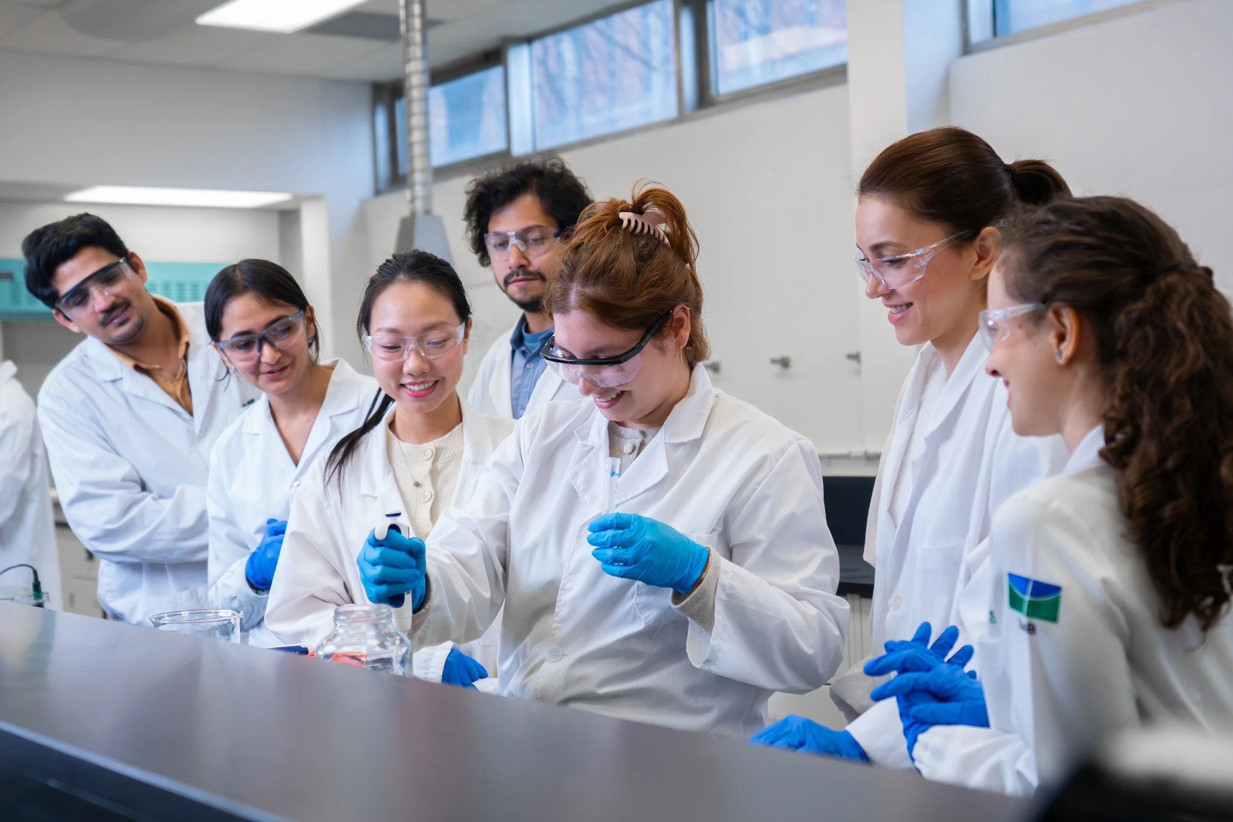Group of scientists wearing white lab coats, safety glasses, and blue gloves working together in a laboratory. They are examining a sample or conducting an experiment.