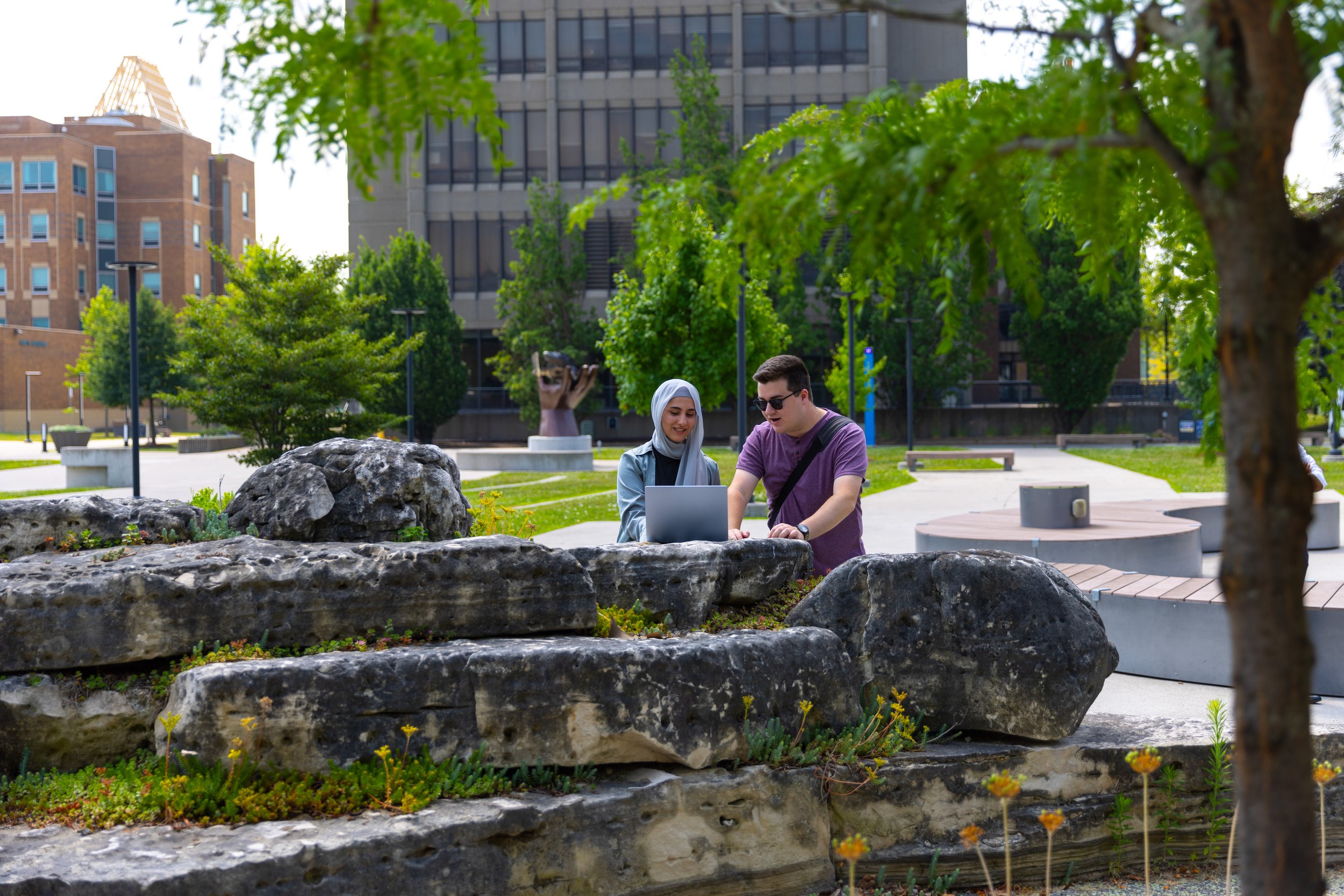 Two people, a woman wearing a hijab and a man wearing sunglasses, working together on a laptop in a park surrounded by trees and rocks, with city buildings in the background.