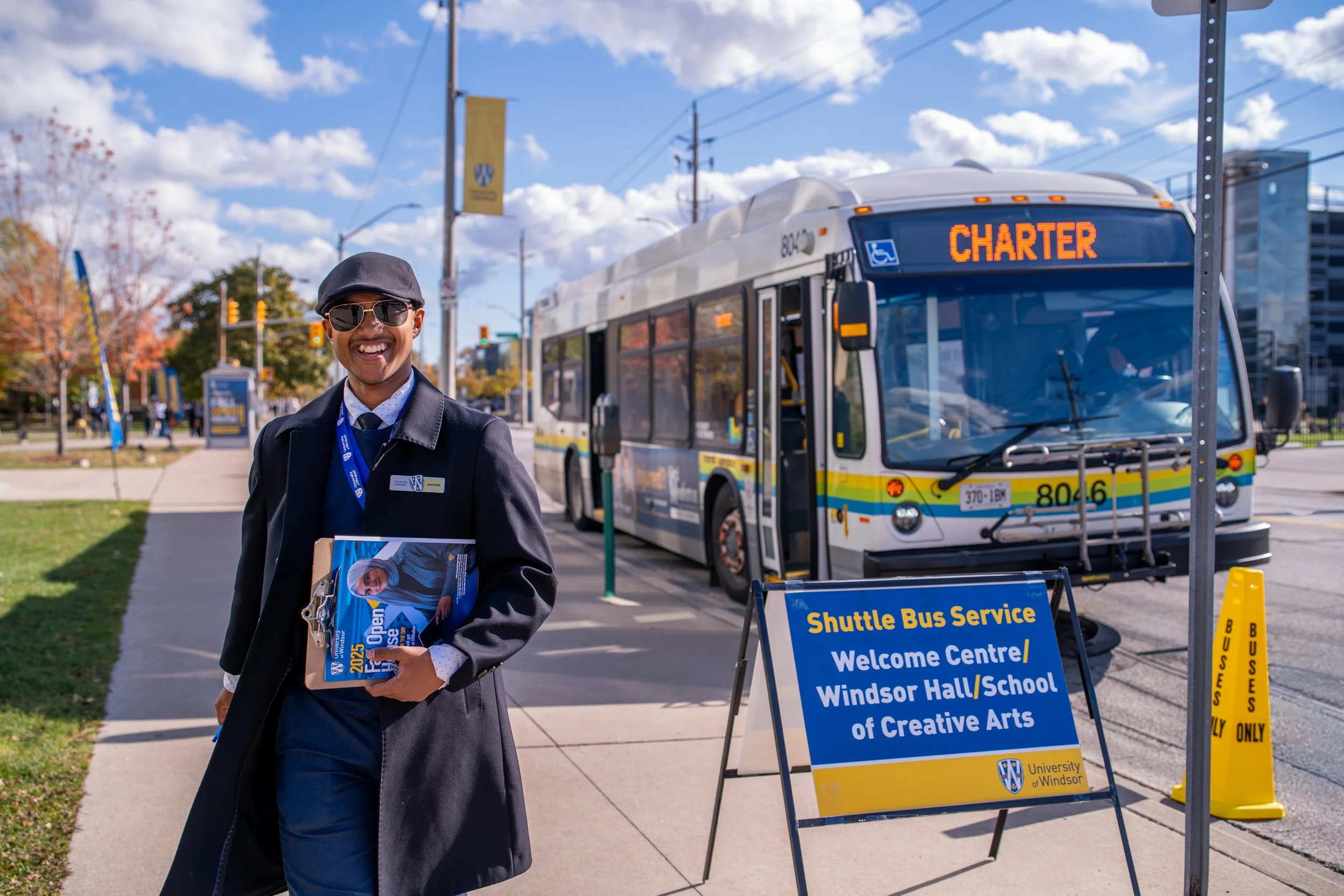A smiling man in sunglasses holding brochures stands next to a shuttle bus with a sign for the University of Windsor. The scene is outdoors on a clear, sunny day with a blue sky, trees, and signs indicating shuttle bus service and a welcome center fo