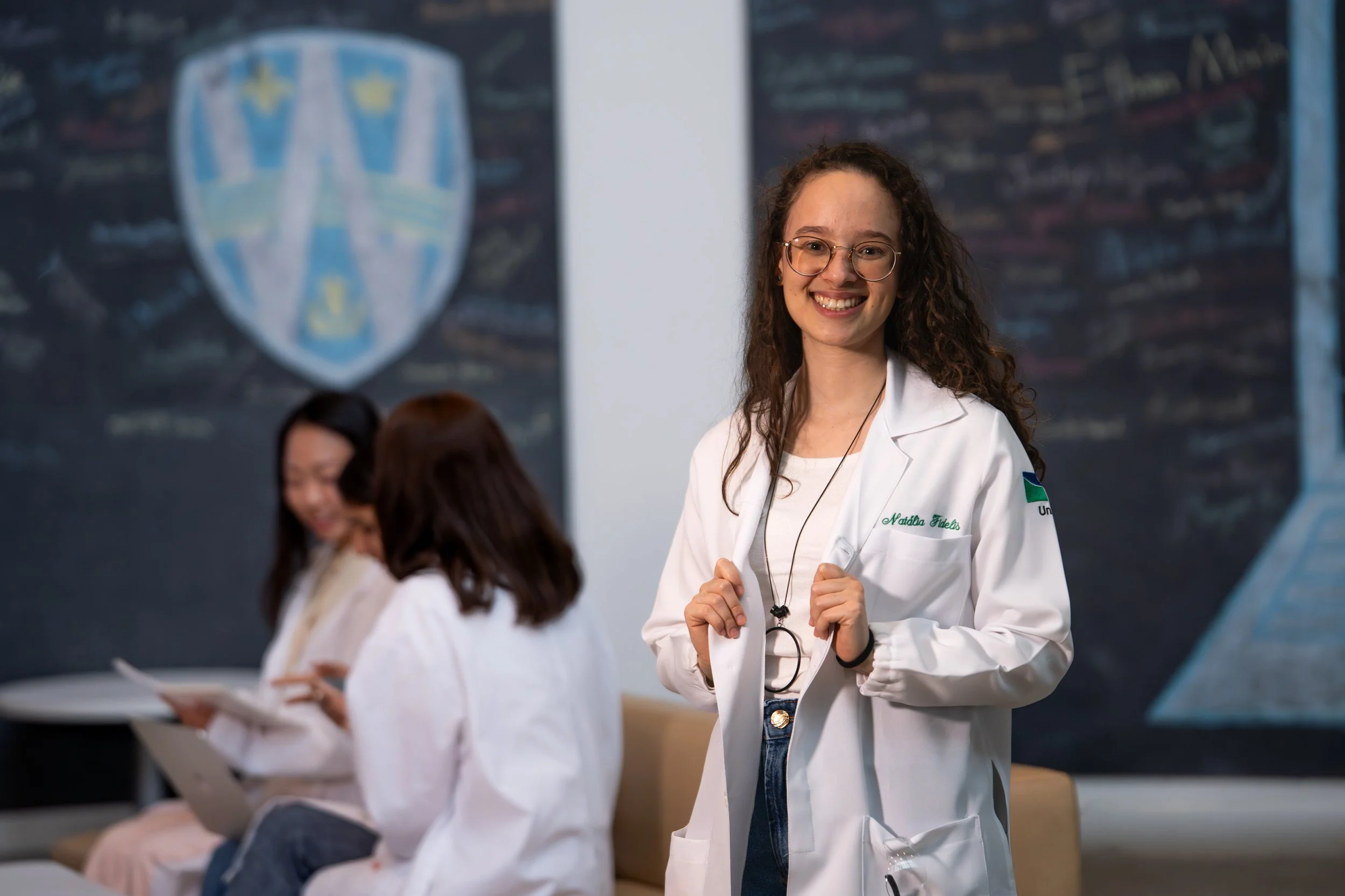 A young woman with curly hair, glasses, and a white lab coat smiling at the camera in a classroom or lab setting, with other students working at desks in the background.