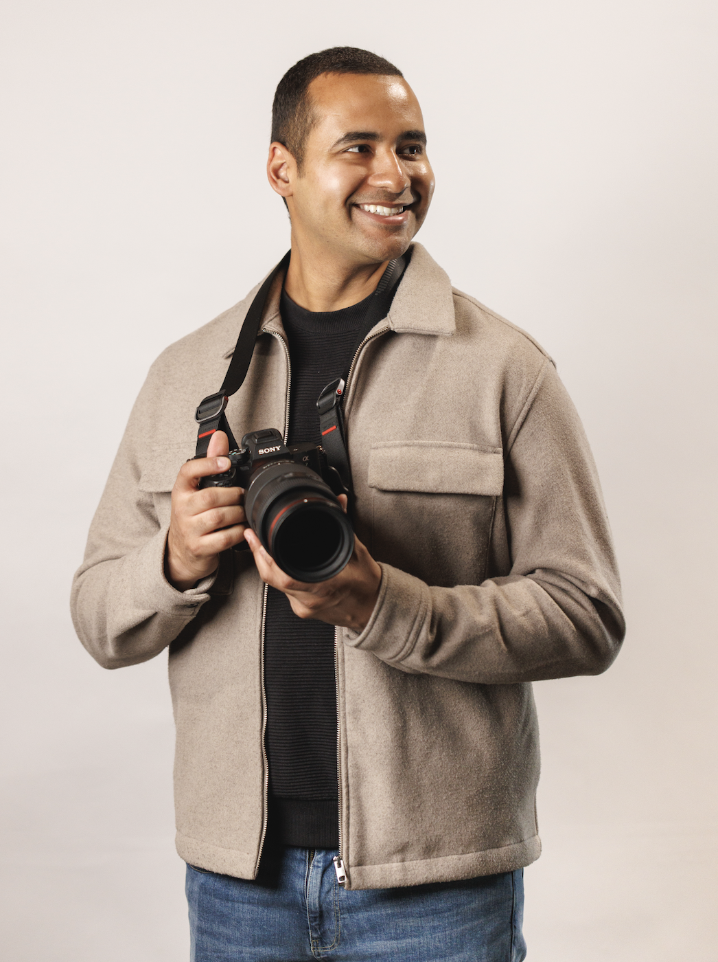 A smiling man holding a Sony camera with a large lens, wearing a beige jacket and a black shirt, standing against a plain white background.