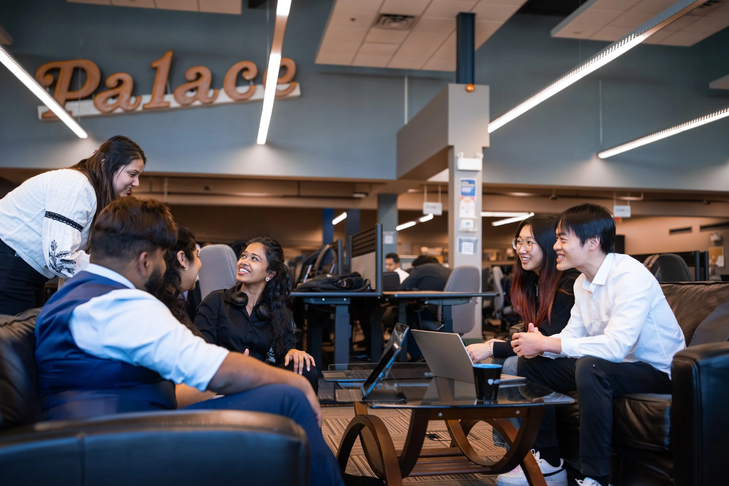Group of young people sitting at tables in a modern office or coworking space, engaging in conversation and smiling, with computers and laptops in front of them. A woman stands smiling and leaning toward the group, behind a blue wall with the word 'P