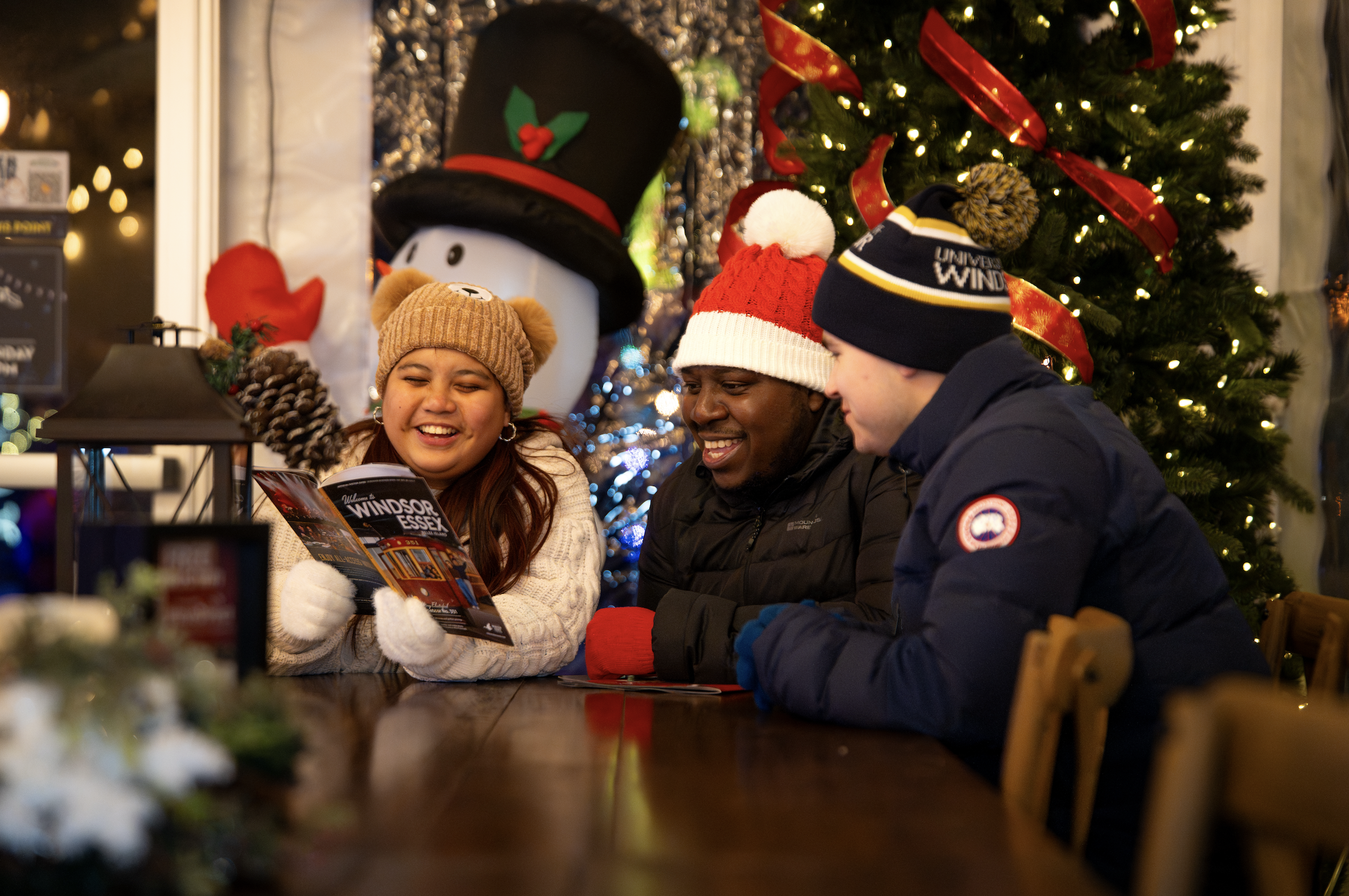 Group of people wearing Christmas hats gathered around a table, smiling and looking at a brochure, with a decorated Christmas tree in the background.
