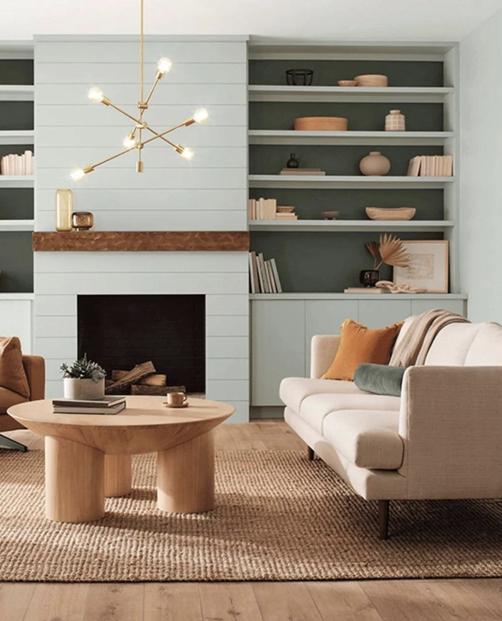 Living room with built-in shelves, a fireplace, a modern chandelier, a beige sofa with pillows, a wooden coffee table with books, and decorative items on the shelves and mantel.