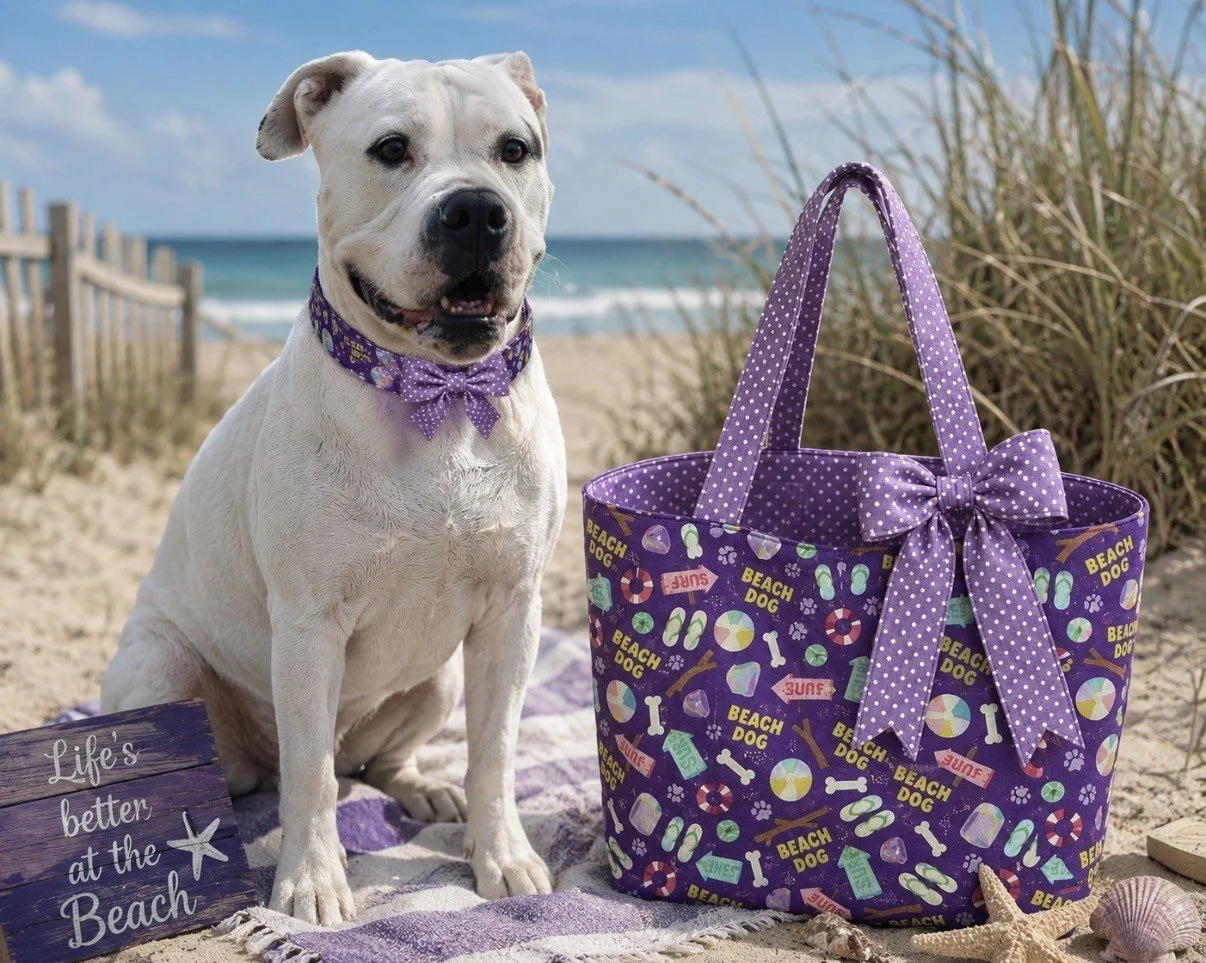 A white dog sitting on a beach blanket next to a purple beach tote with polka dot bow, decorated with summer and beach-themed illustrations, and a lavender polka dot collar with bow. The background shows sand, tall grass, ocean, and blue sky.