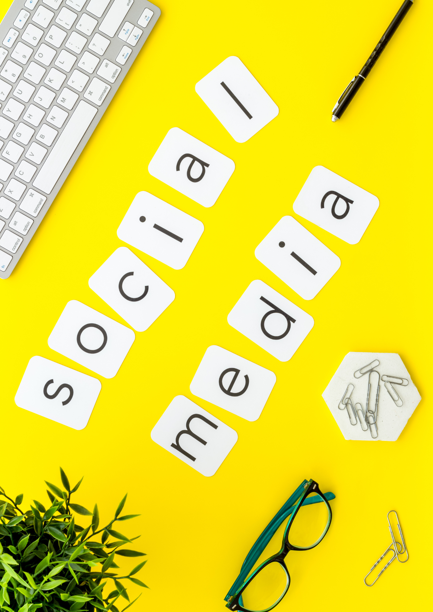 Office desk setup with scattered letter tiles spelling 'email' on a yellow surface, a computer keyboard, a pen, a pair of teal glasses, green plants, and paper clips.