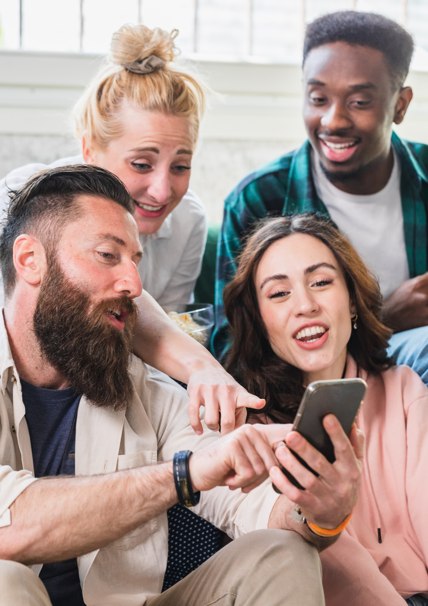 Group of five friends looking at a smartphone together, sitting indoors near a window, smiling and enjoying each other's company.