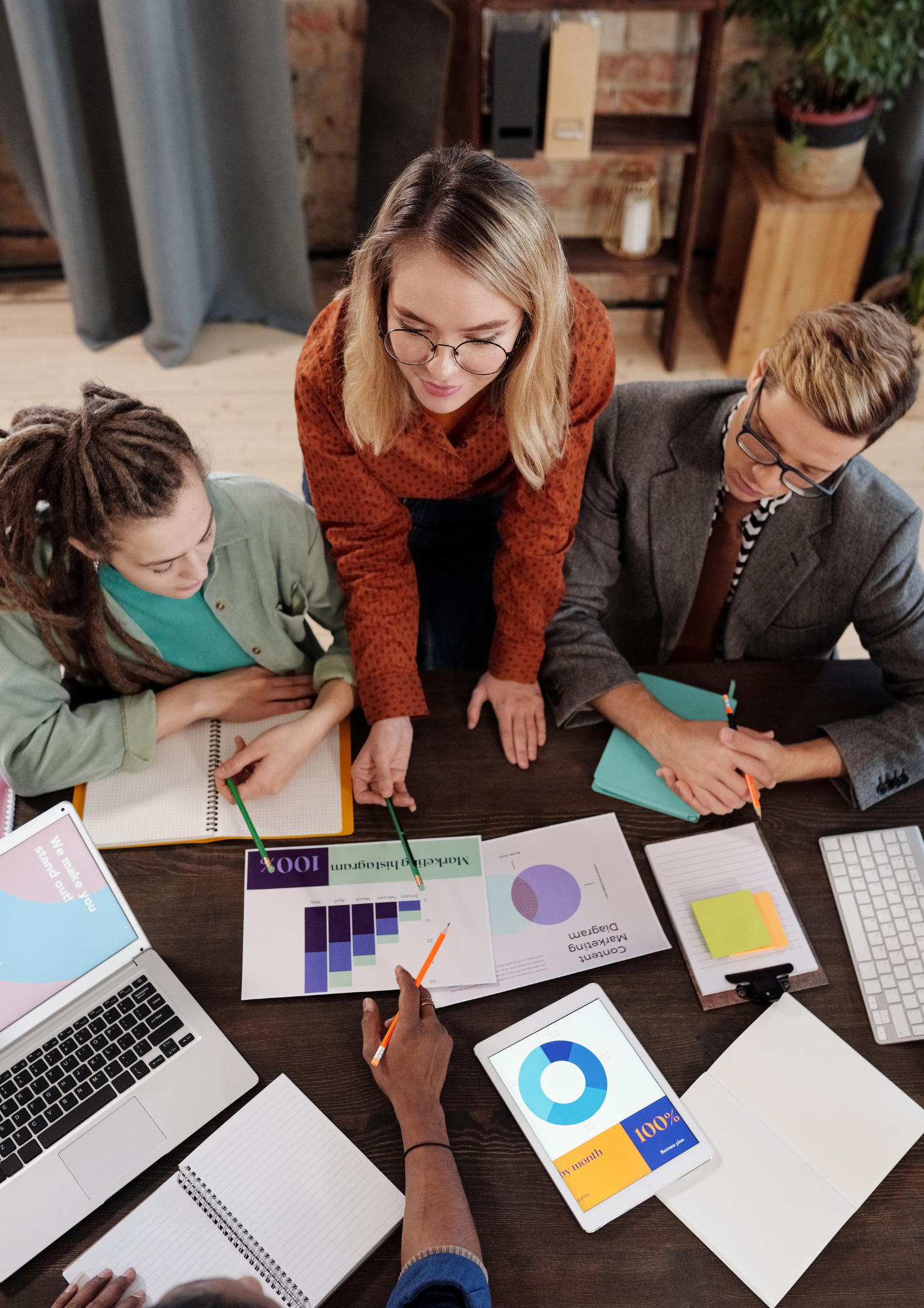 A group of people in a meeting discussing data and charts with laptops, tablets, notebooks, and printed graphs on the table.