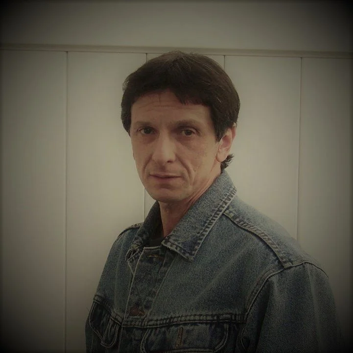 A man with dark hair wearing a denim jacket standing in front of a plain wall.