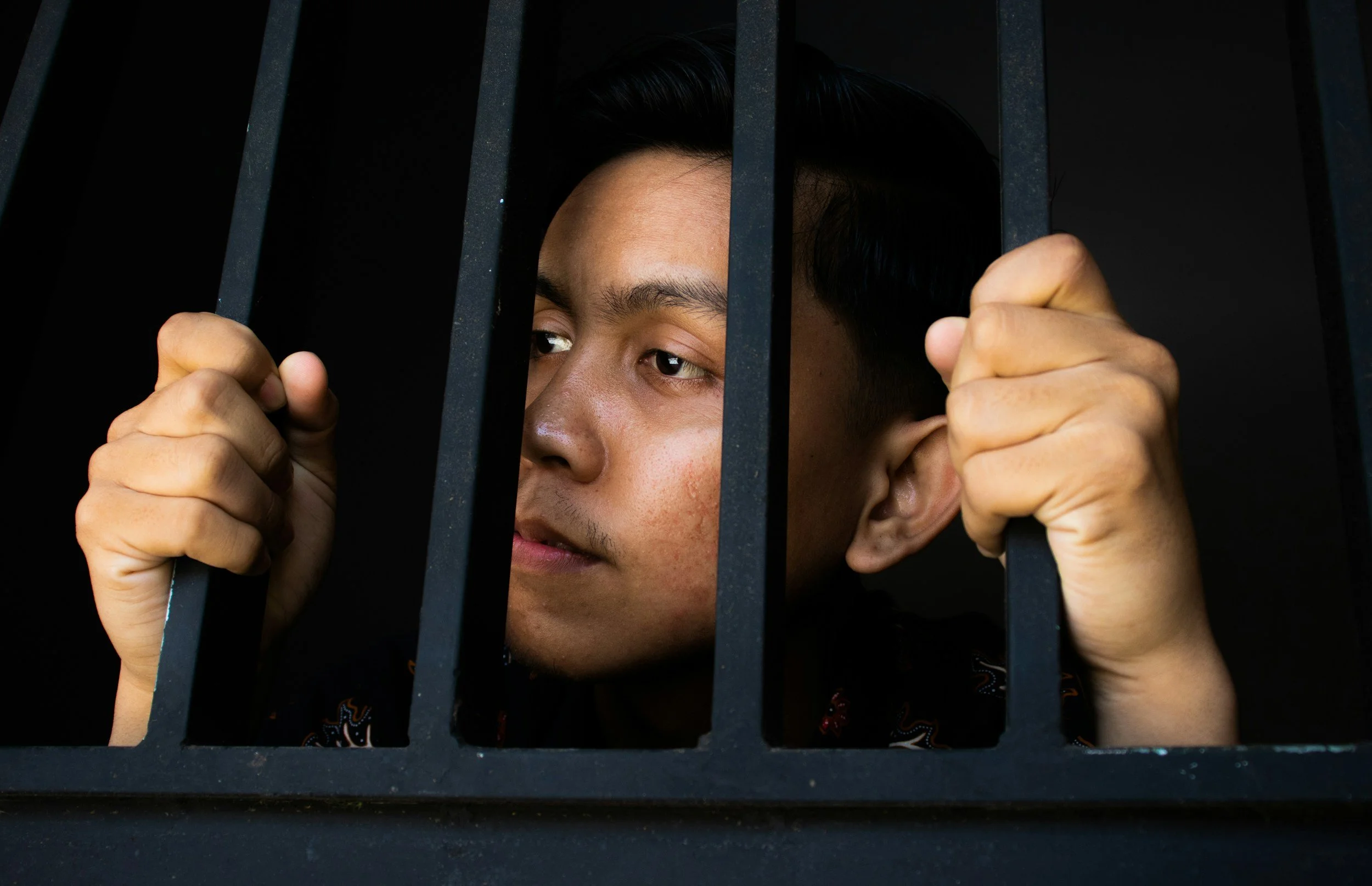 Person looking through bars, holding the bars with both hands, dark background.