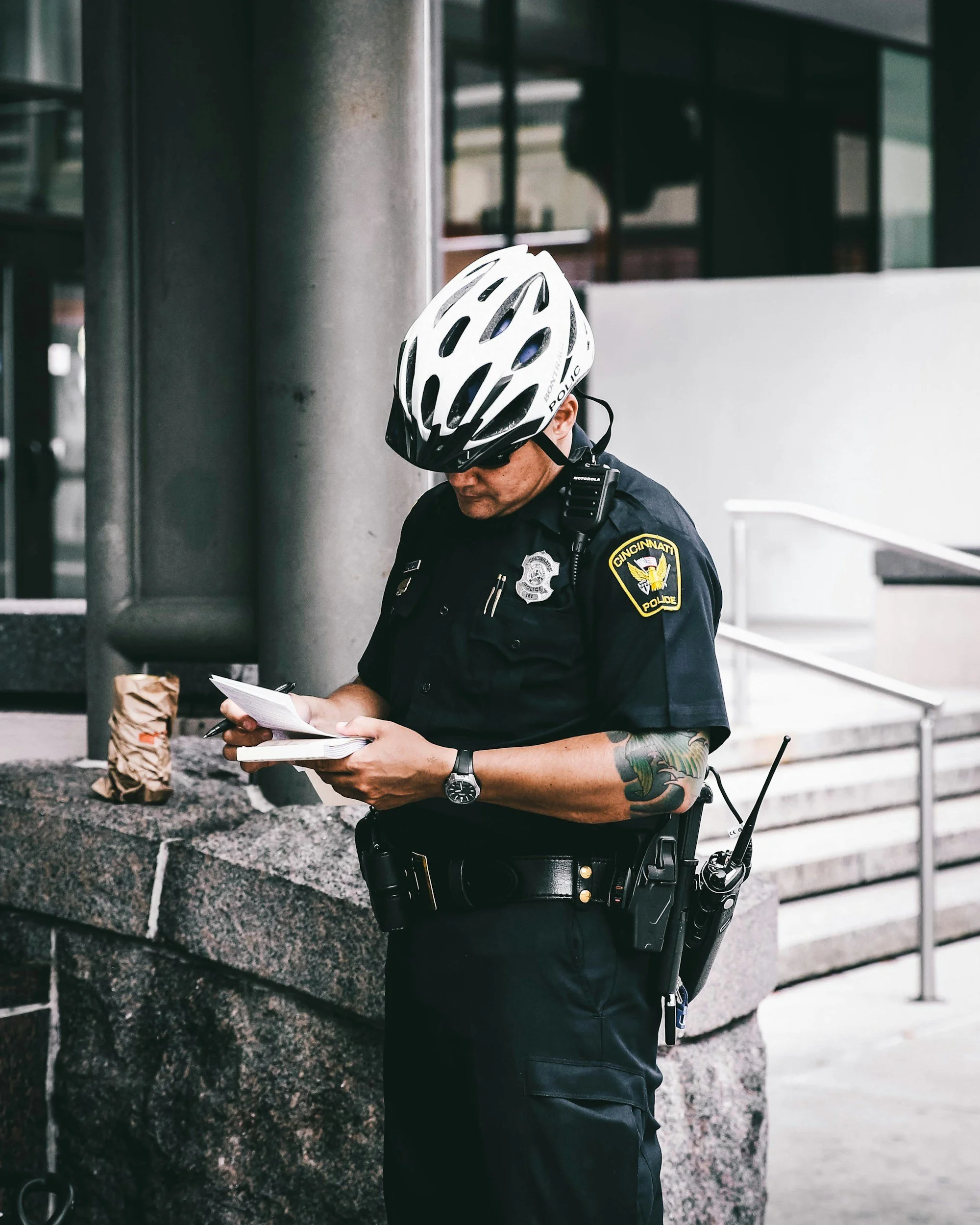 A police officer wearing a white helmet and sunglasses is standing near a stone building, reading a notepad. The officer has colorful tattoos on their left arm and wears a police badge and equipment on their uniform.