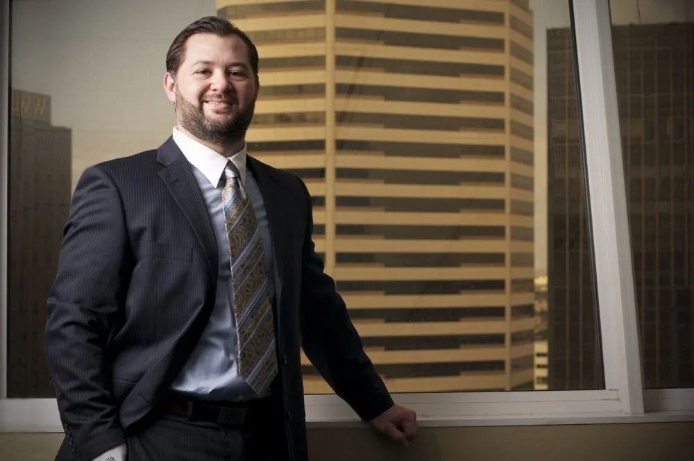 A man in a suit and tie standing by a window in an office building, with city skyscrapers in the background.