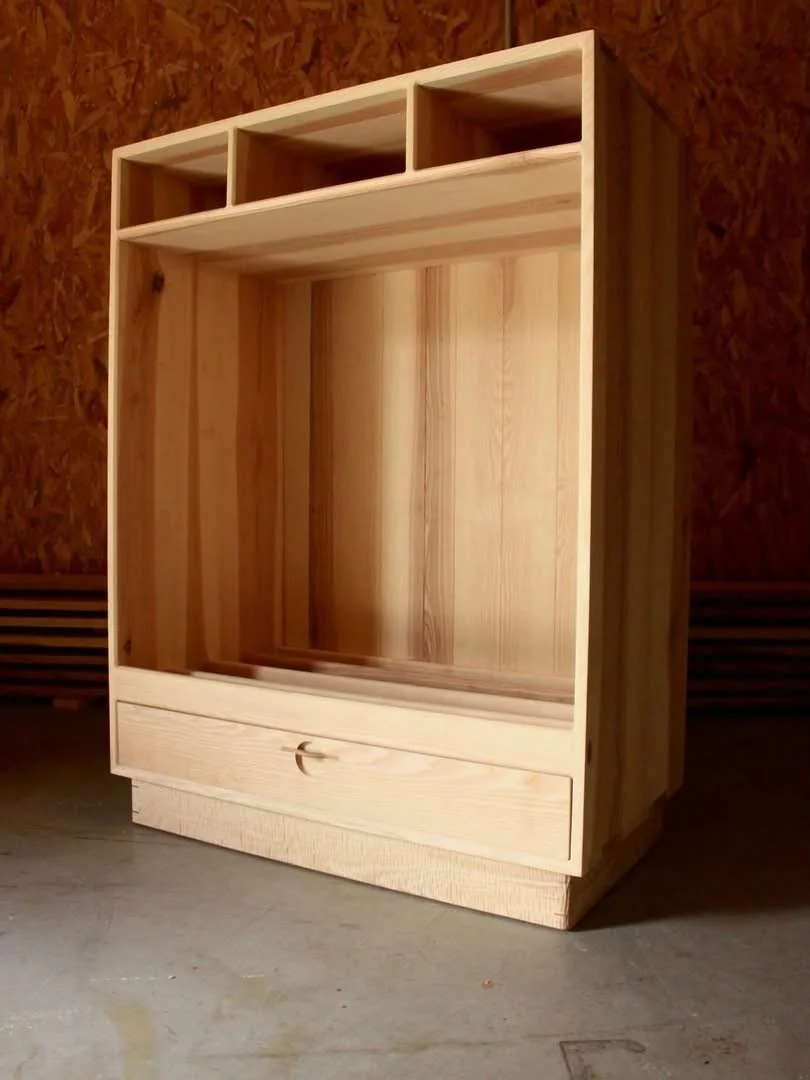 Une étagère en bois clair avec plusieurs compartiments, y compris une grande section au centre, une petite trappe en dessous, et trois petits tiroirs en haut, placée contre un mur en bois.