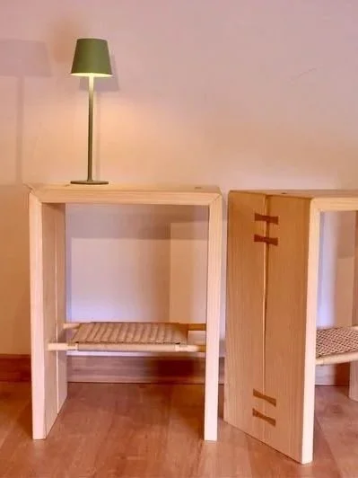 Deux tables en bois clair avec une lampe verte posée sur la première. La première table a une étagère en dessous, tandis que la deuxième est retournée, montrant ses côtés avec des détails en bois.