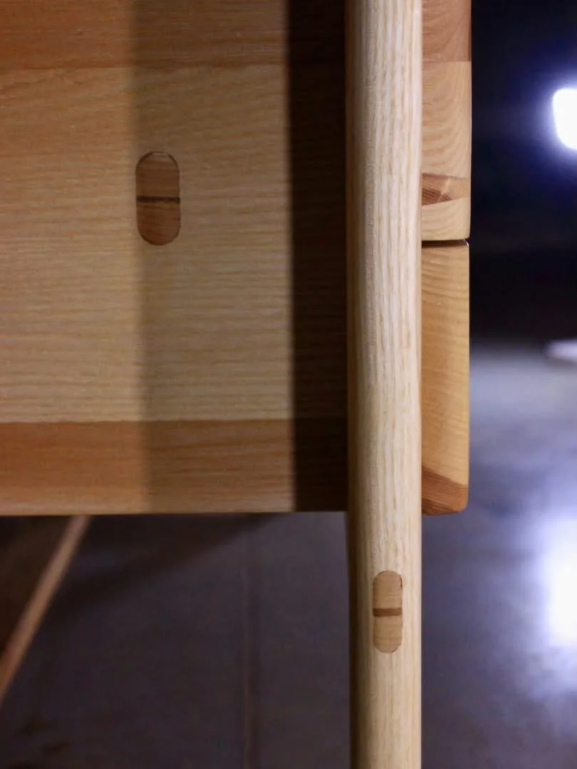 Détail d'un meuble en bois avec des pièces connectées à tenons morts, mettant en évidence la construction en bois. On voit une partie d'une table ou d'une étagère en bois clair avec une pièce verticale qui s'insère dans une ouverture.