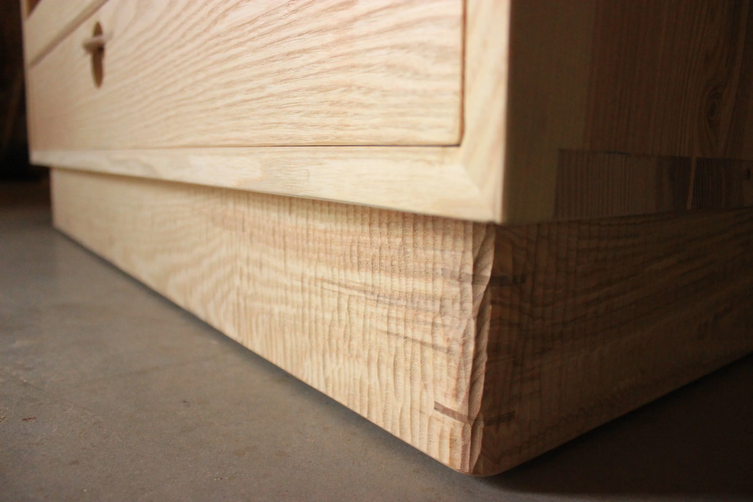 Une pièce de bois brute avec des bords légèrement arrondis, posé sur un sol en béton. Visible grain de bois clair et texture naturelle.