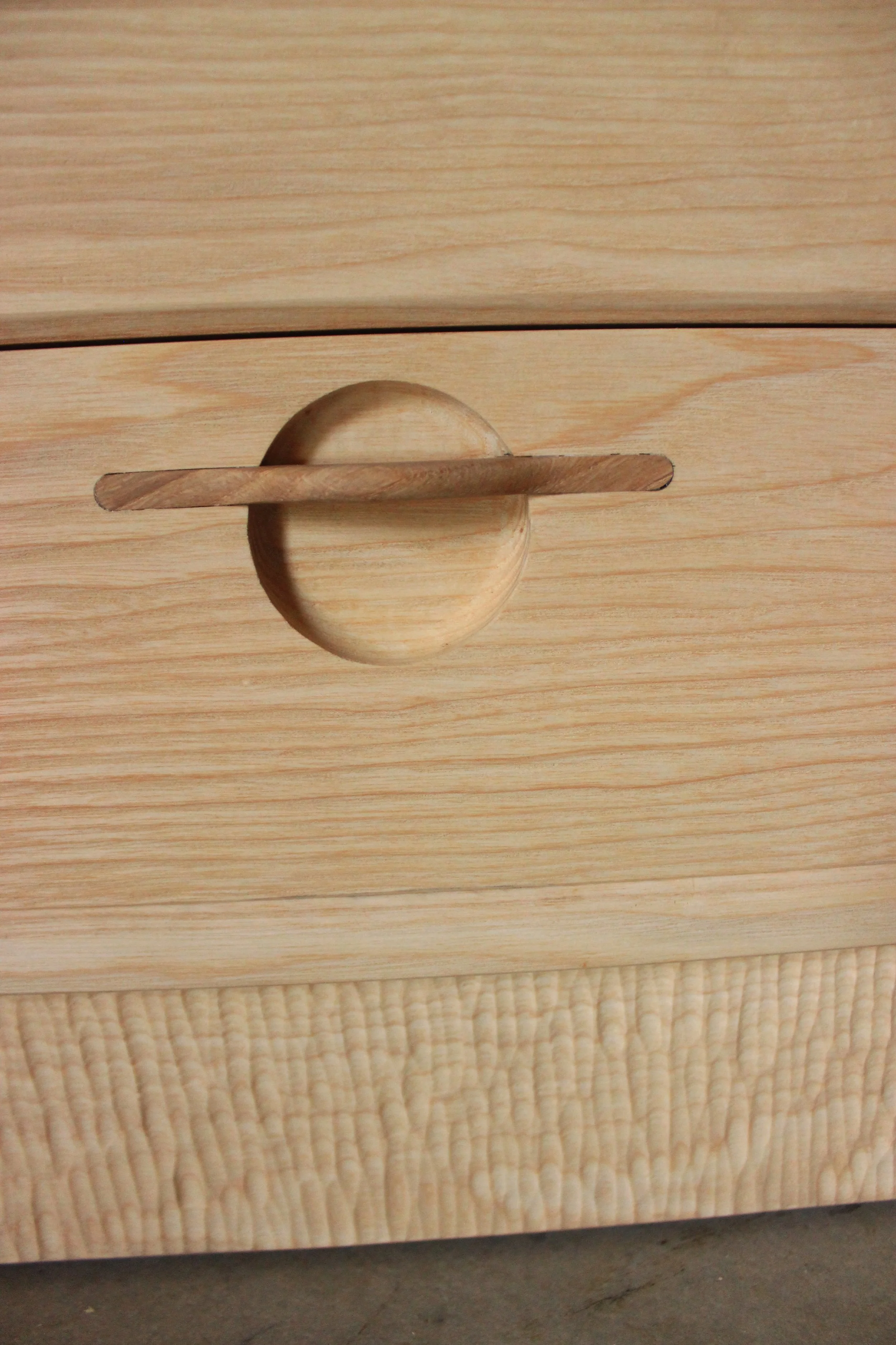 Vue d'une commode en bois clair avec une poignée ronde en bois, placée sur un tapis beige. Le fond ne montre que le sol en béton.