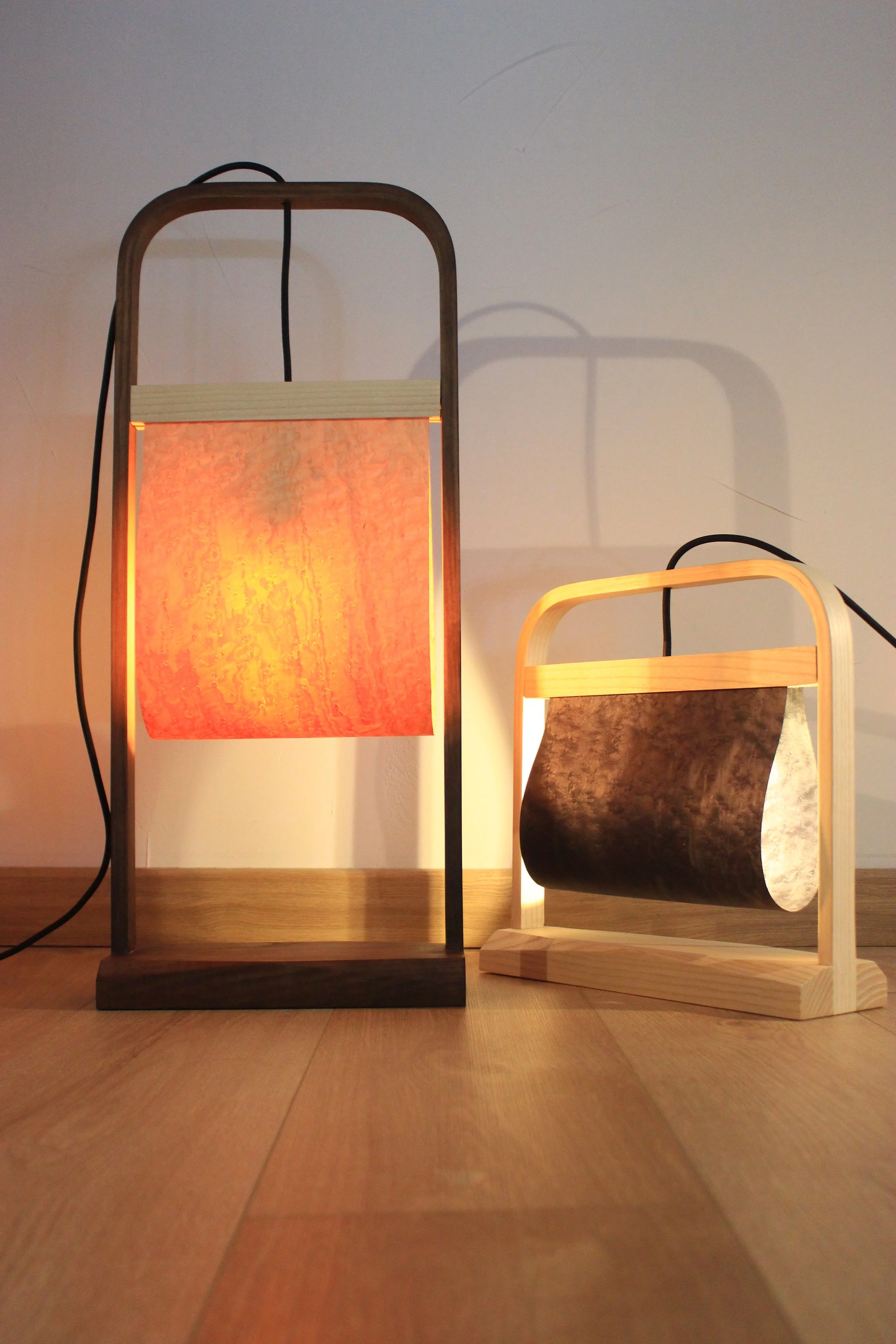 Deux lampes en bois, l'une avec un abat-jour orange et l'autre avec une ombre en bois sculpté, sont placées sur un sol en bois contre un mur blanc.