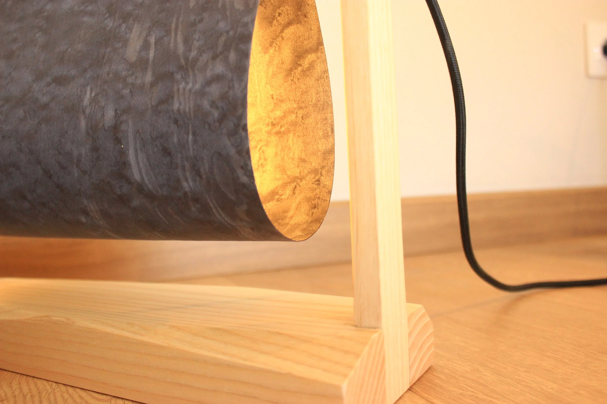 Une lampe de table avec une base en bois clair, un abat-jour en métal sombre, et un cordon électrique noir, posée sur une surface en bois clair.