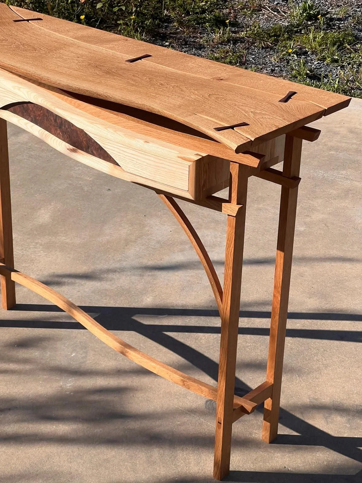 Une table en bois avec un design unique, alliant différentes essences de bois, située à l'extérieur sur un sol en béton, avec des herbes et de la végétation à l'arrière-plan.