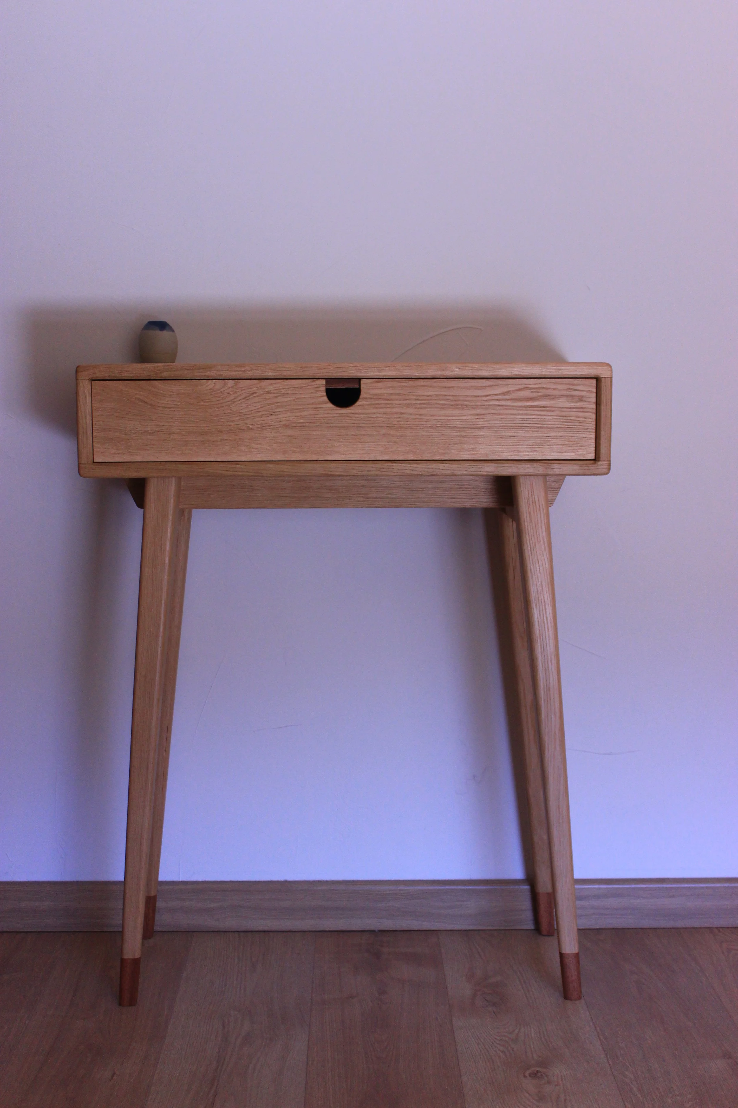 Un meuble en bois clair avec une seule tiroir, soutenu par quatre jambes, placé contre un mur blanc, avec un vase à côté sur le côté gauche.