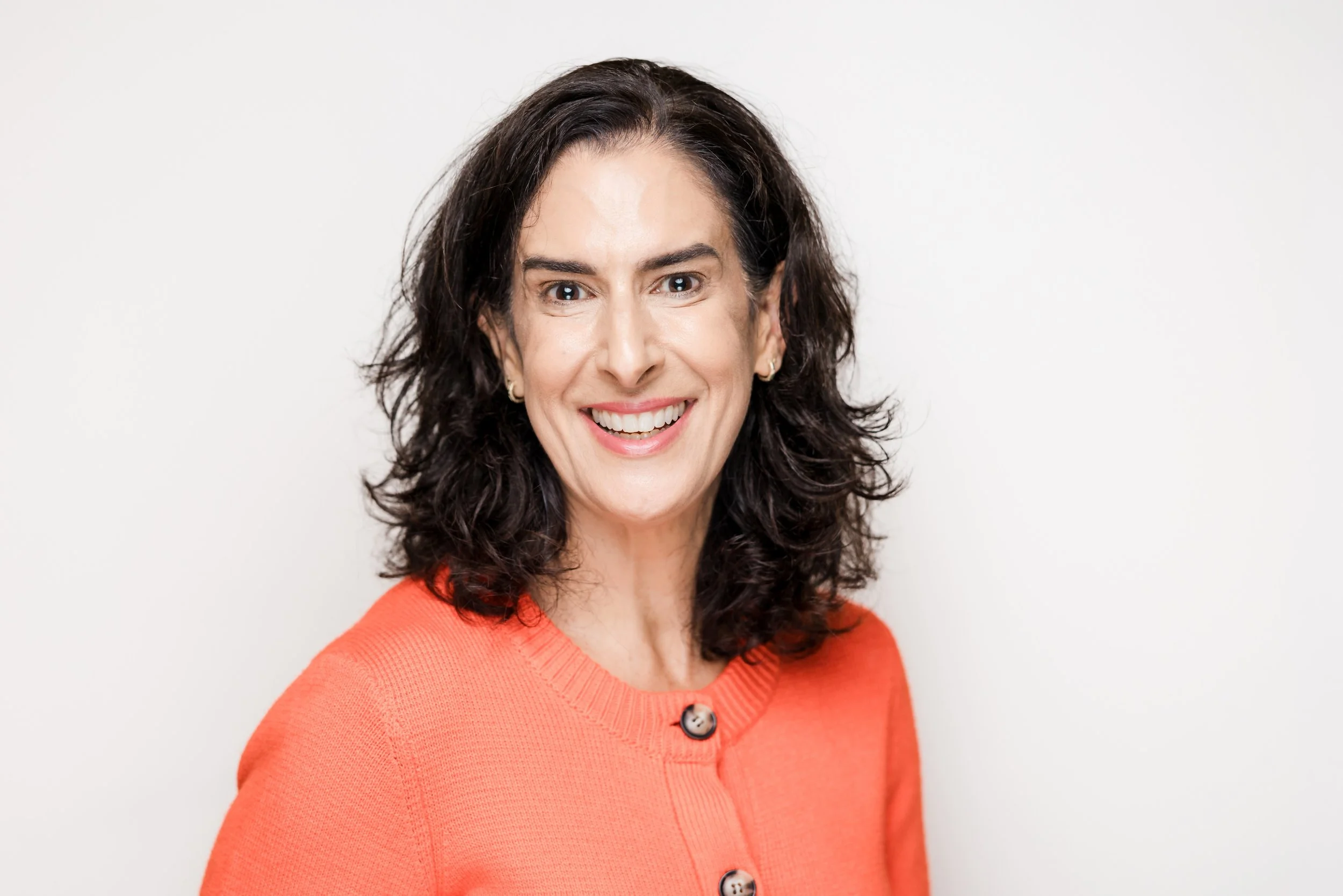 A smiling woman with dark, curly hair wearing an orange sweater with buttons, standing against a white background.