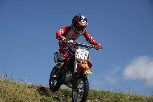 A person in red and black motocross gear riding a dirt bike downhill on a grassy hill under a blue sky.