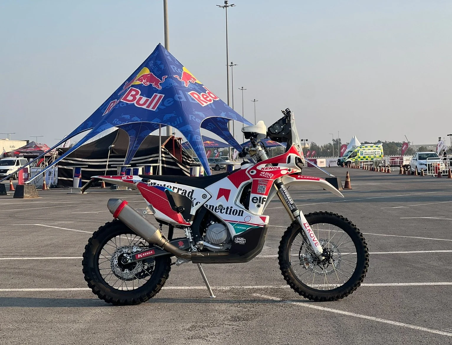 Motorcycle parked in an open area with a Red Bull tent and other vehicles and tents in the background.