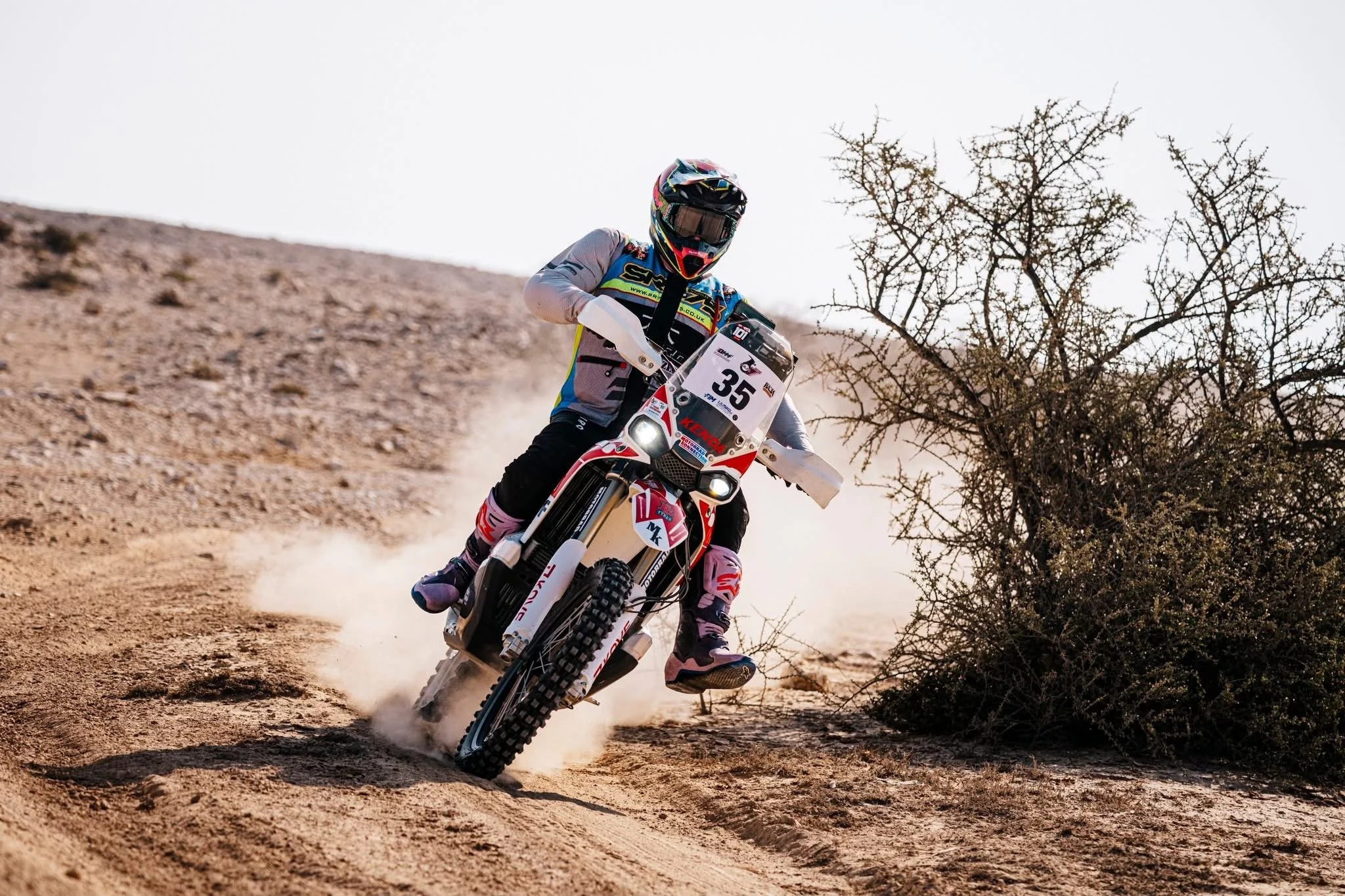 Motocross rider in full gear racing on a dirt trail in a desert landscape with scattered bushes and a clear sky.