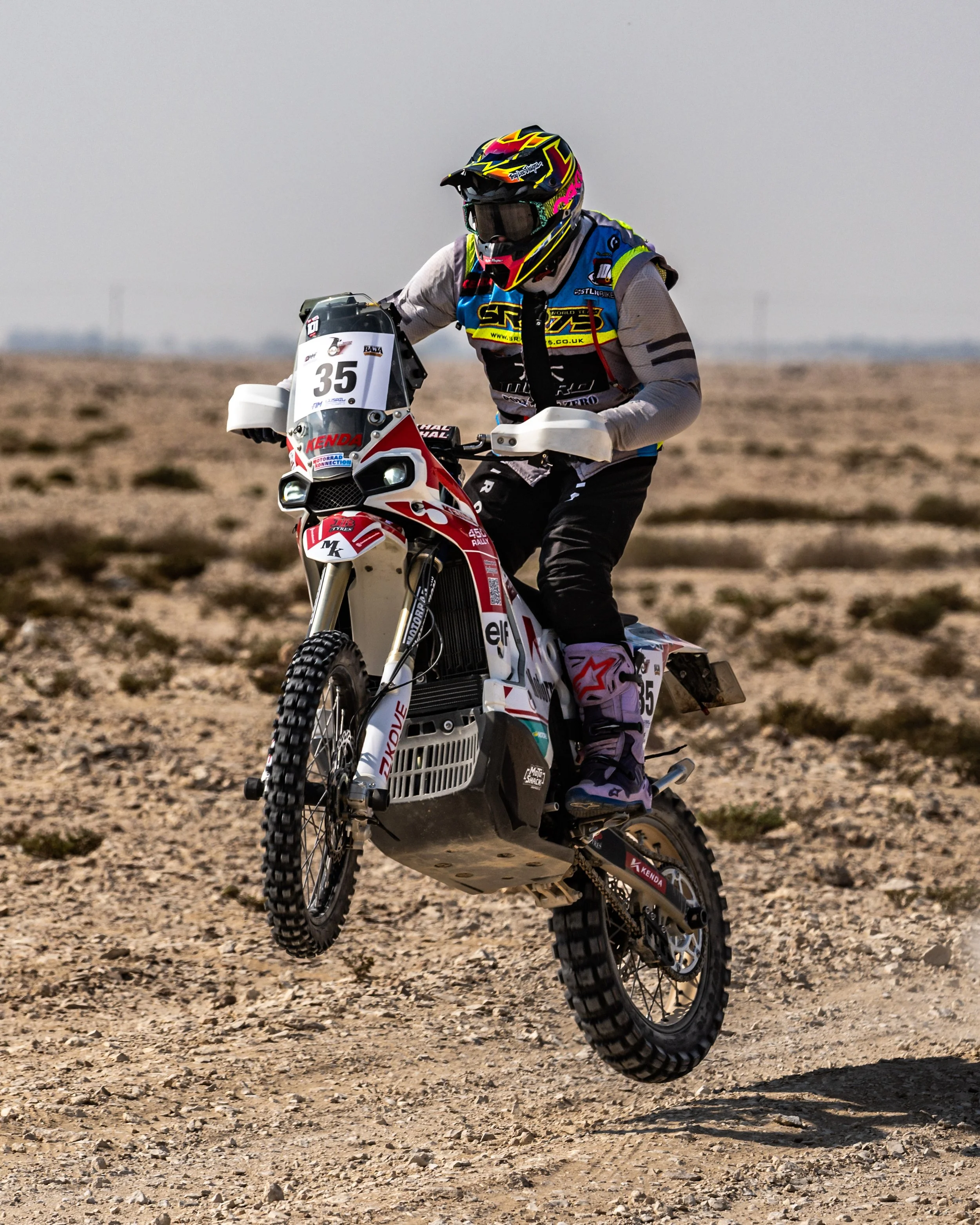 A person riding a rally motorcycle on a rocky, desert landscape, wearing a colorful helmet and protective gear.