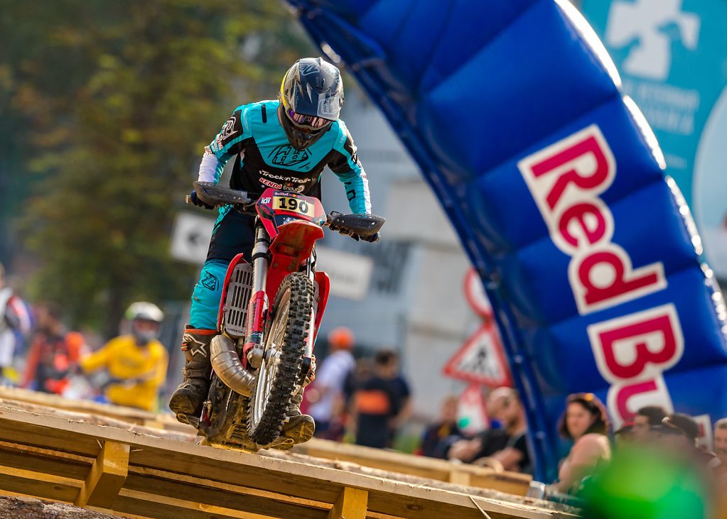 A mountain biker wearing a blue jersey and helmet is riding a red bike over a wooden ramp during a race, with spectators and a Red Bull arch in the background.