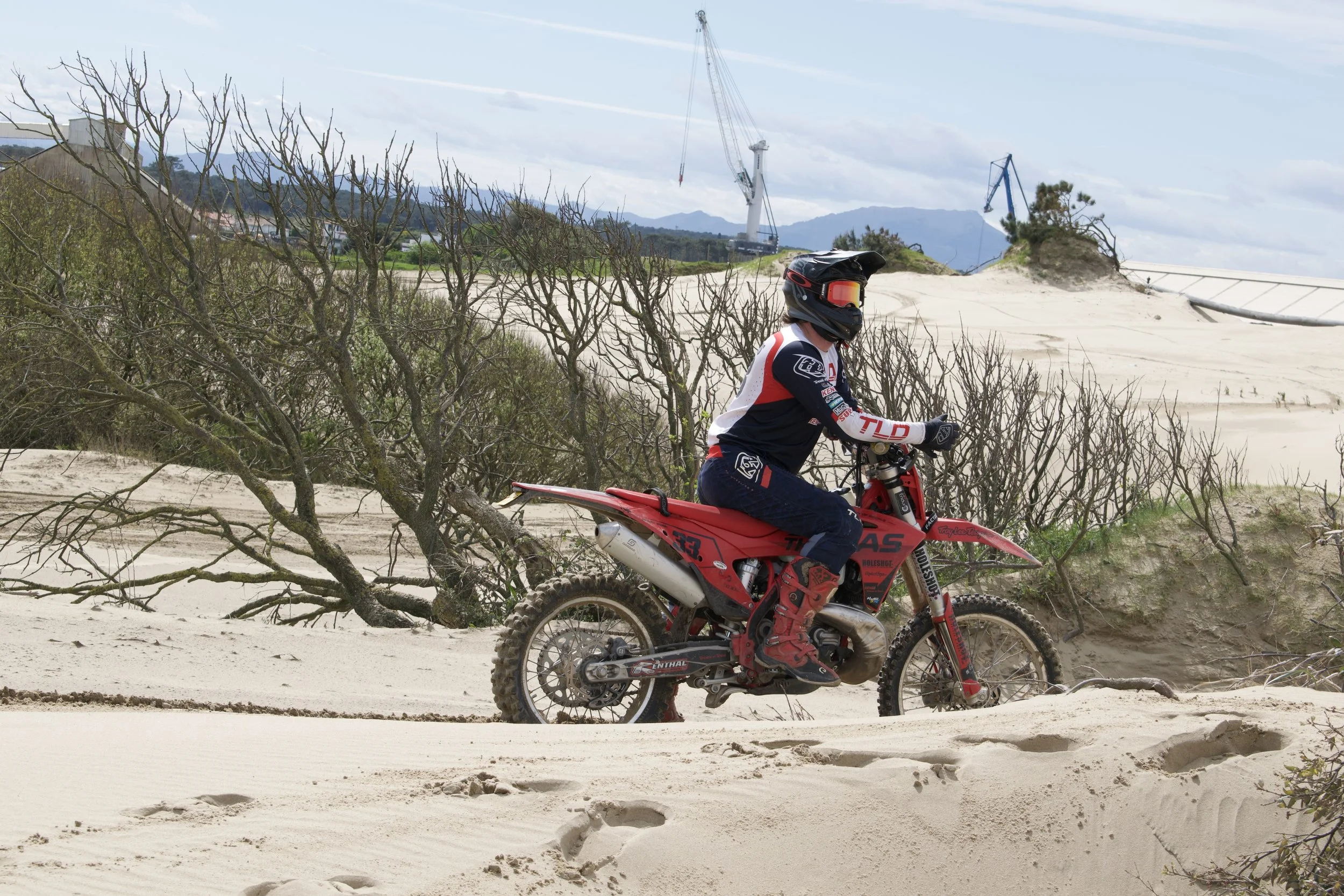 A person wearing a black helmet, goggles, and motocross gear riding a red dirt bike on sandy terrain near some leafless bushes, with construction cranes and distant hills in the background.
