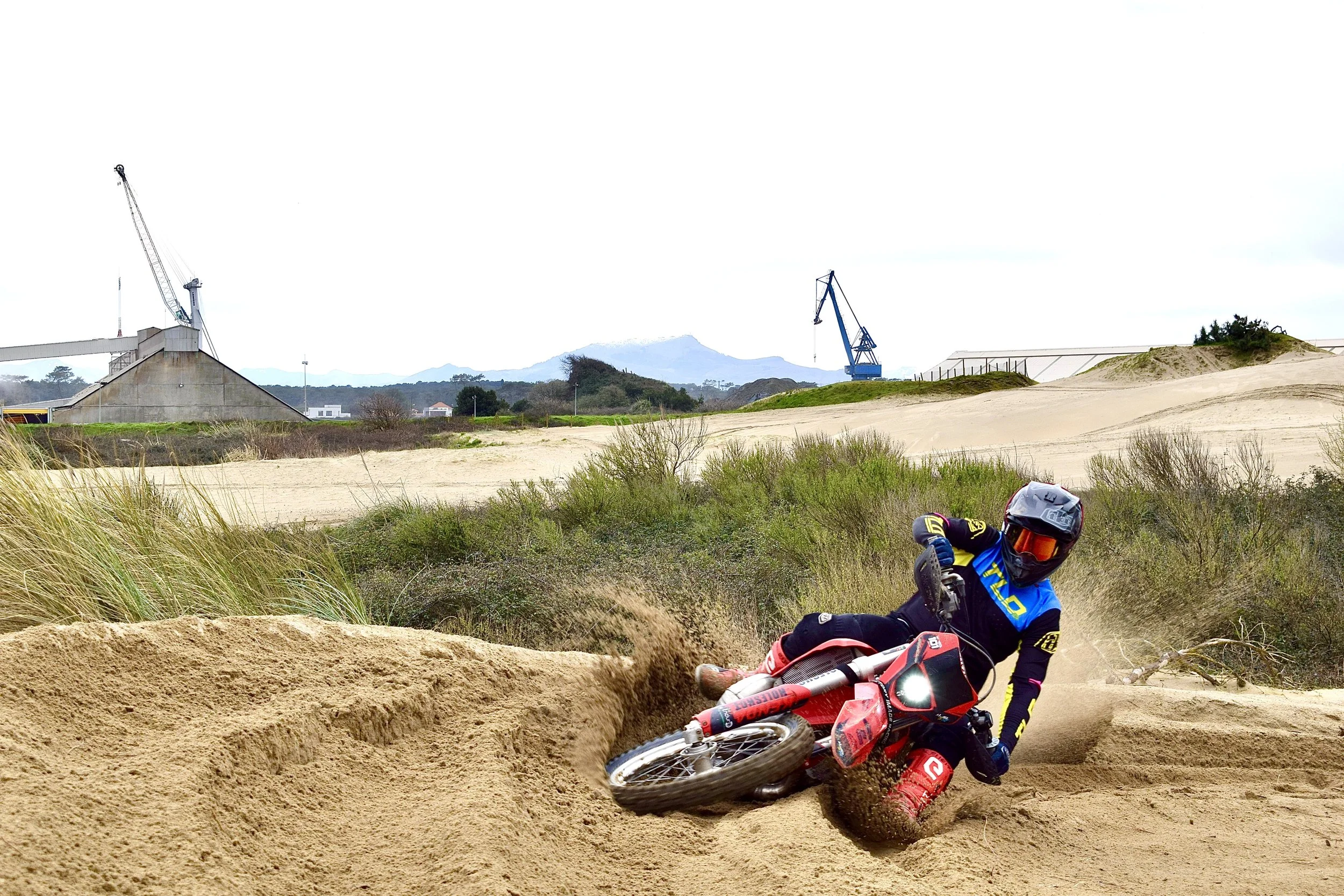 A motocross rider crashes on a dirt track, falling off and wobbling the motorcycle while wearing a blue and yellow racing suit and helmet, with dirt flying around.