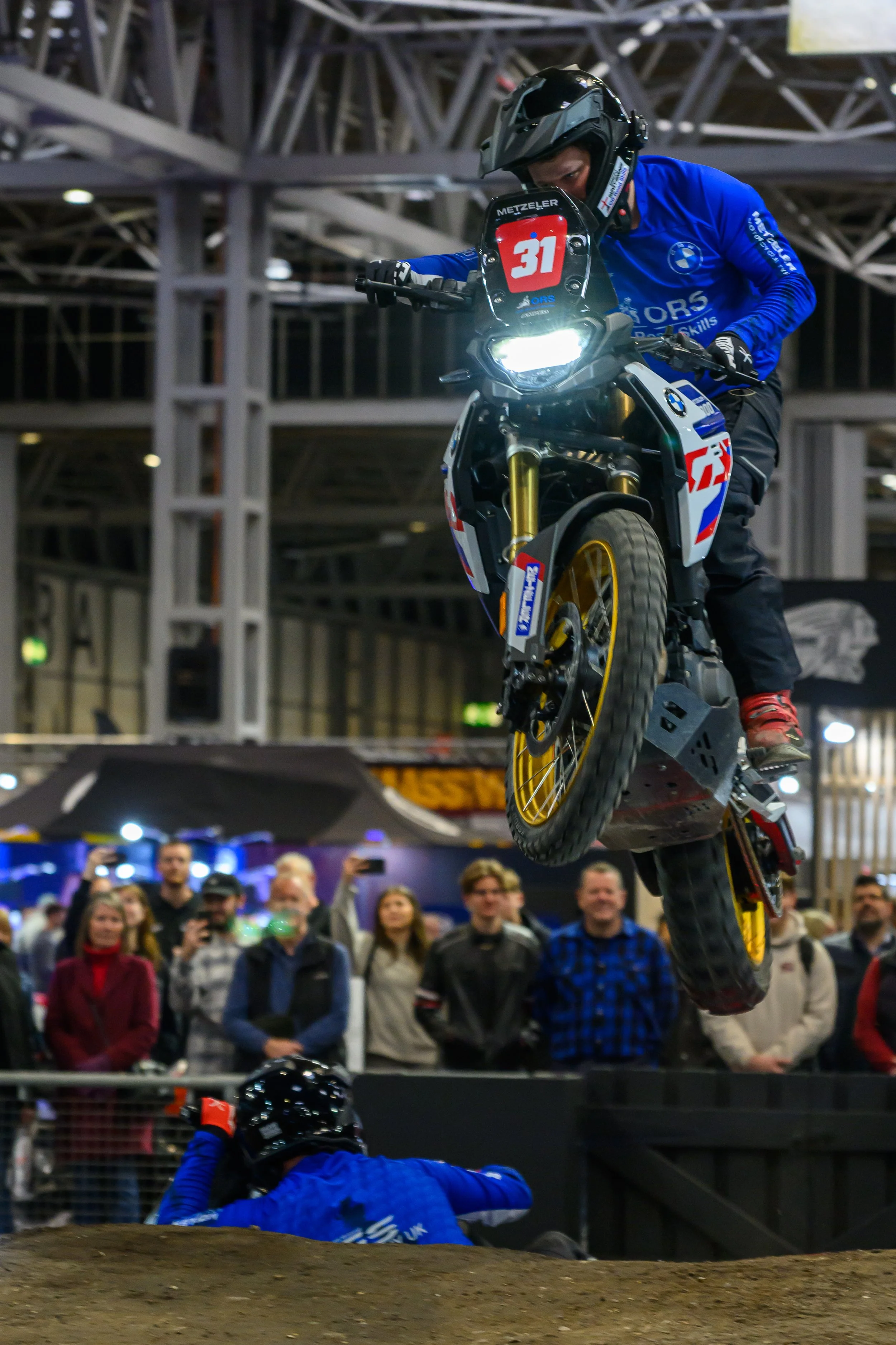 Motocross rider performing a trick in mid-air at an indoor event, with a crowd watching below.