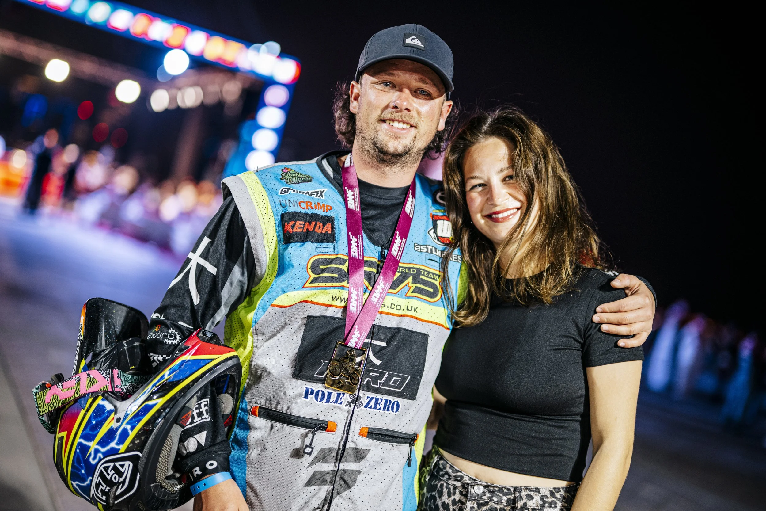 A man in motocross gear smiling, with a woman beside him embracing him, at night with blurred bright lights in the background.
