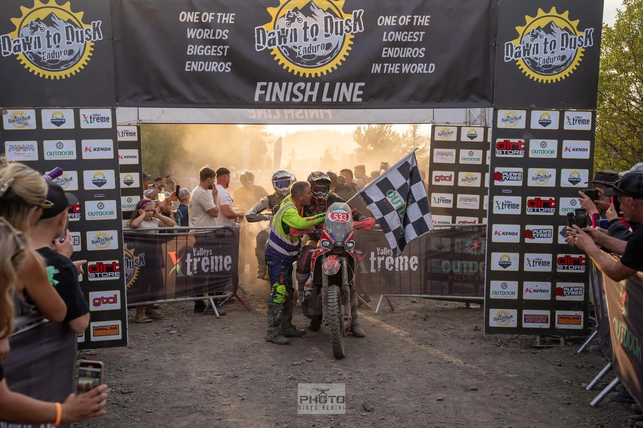 Motocross rider in full gear crossing the finish line at an off-road endurance race, with spectators on either side taking photos. The backdrop features banners with race and sponsor logos, and the setting sun creates a dusty haze.