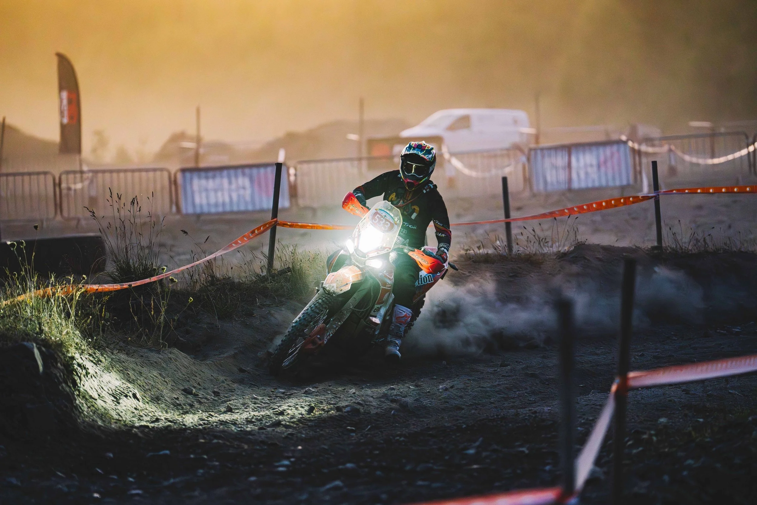 An off-road motorcycle rider wearing a helmet and gear navigating a dirt track at sunset, with dust and a fence in the background.