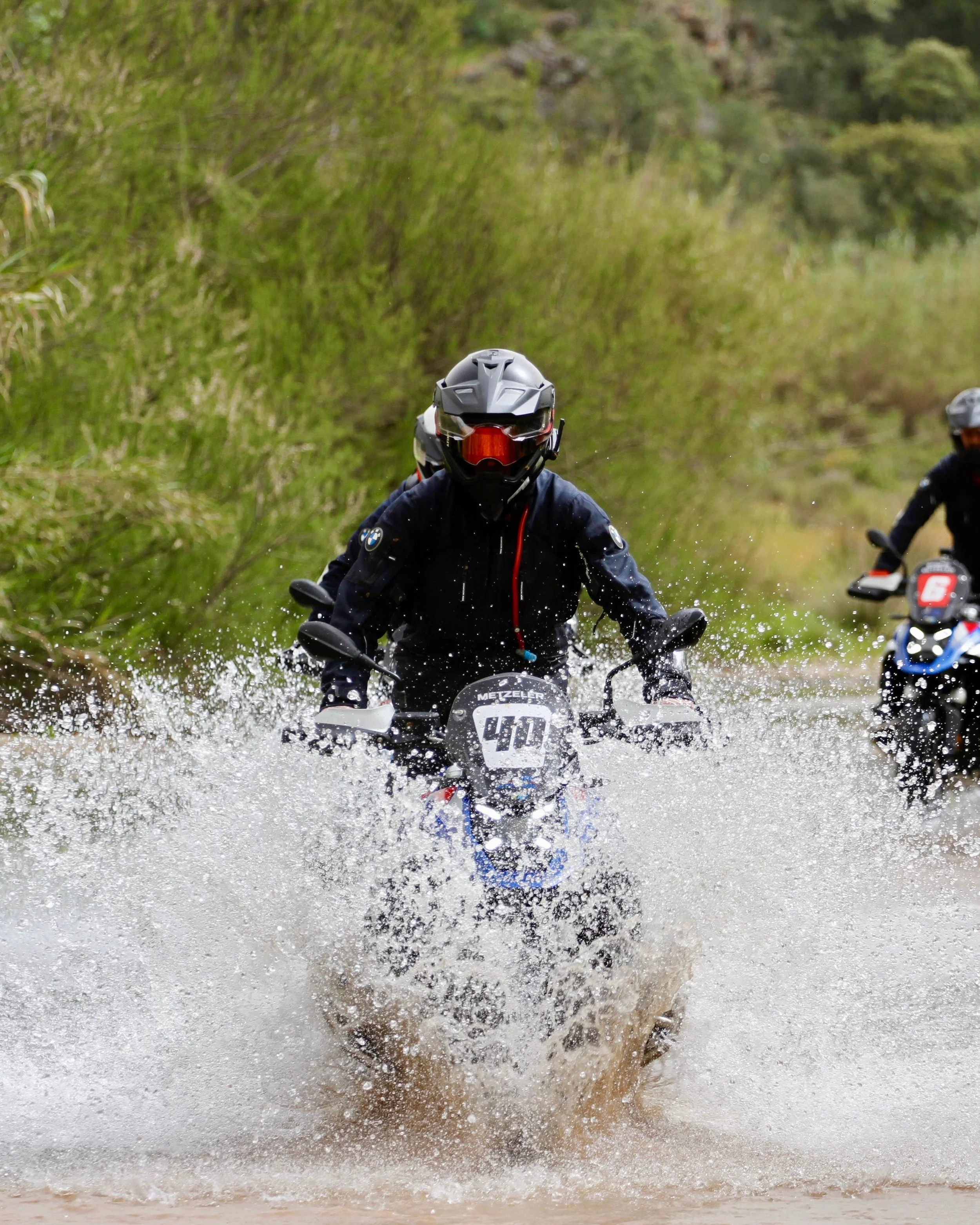 Motorcyclists riding through a shallow water crossing in a lush green outdoor setting.