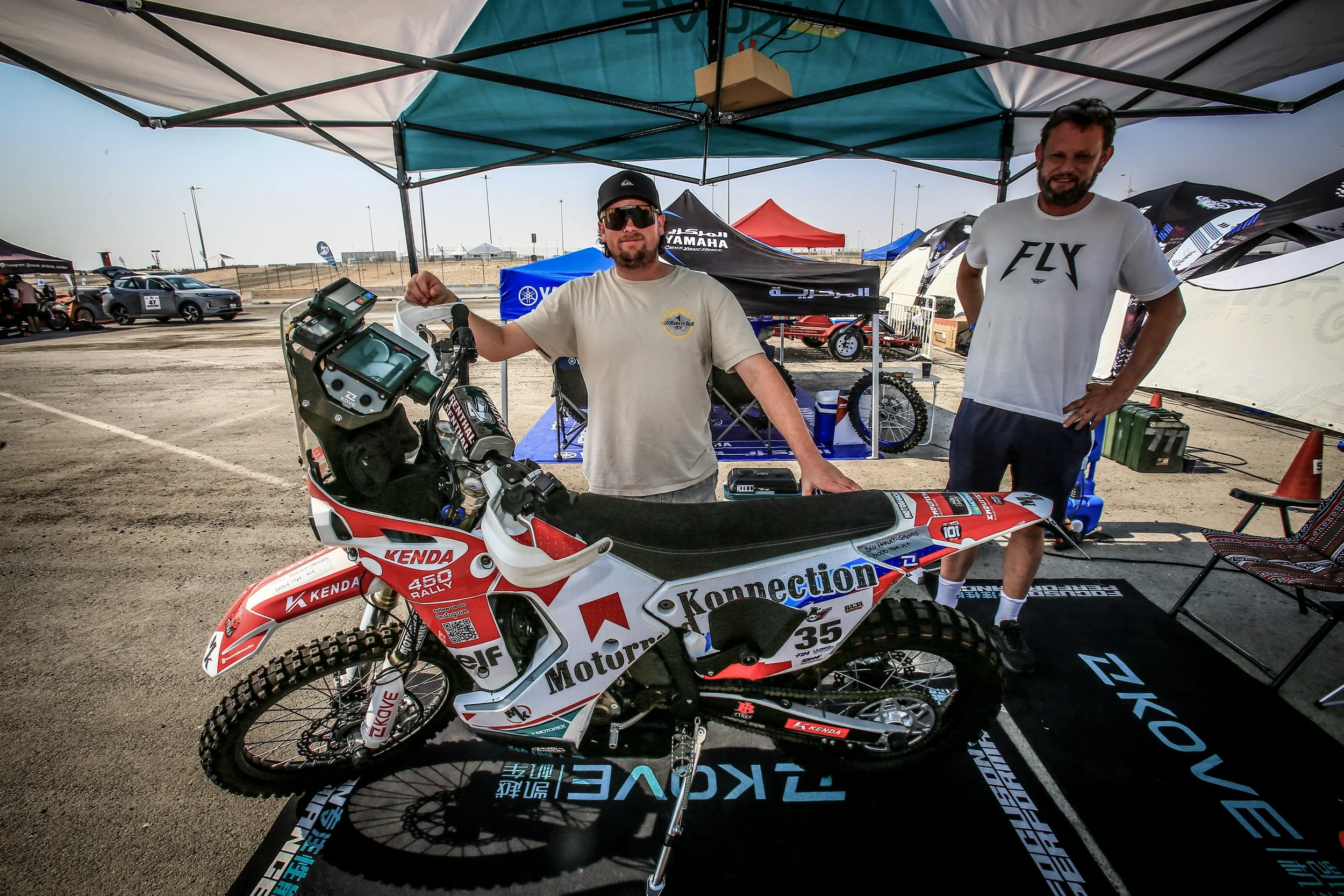 Two men stand under a canopy tent behind a rally motorcycle at an outdoor race event. The motorcycle is decorated with sponsor logos and the numbers 35 and 450. In the background, there are other tents, vehicles, and a dirt race track.