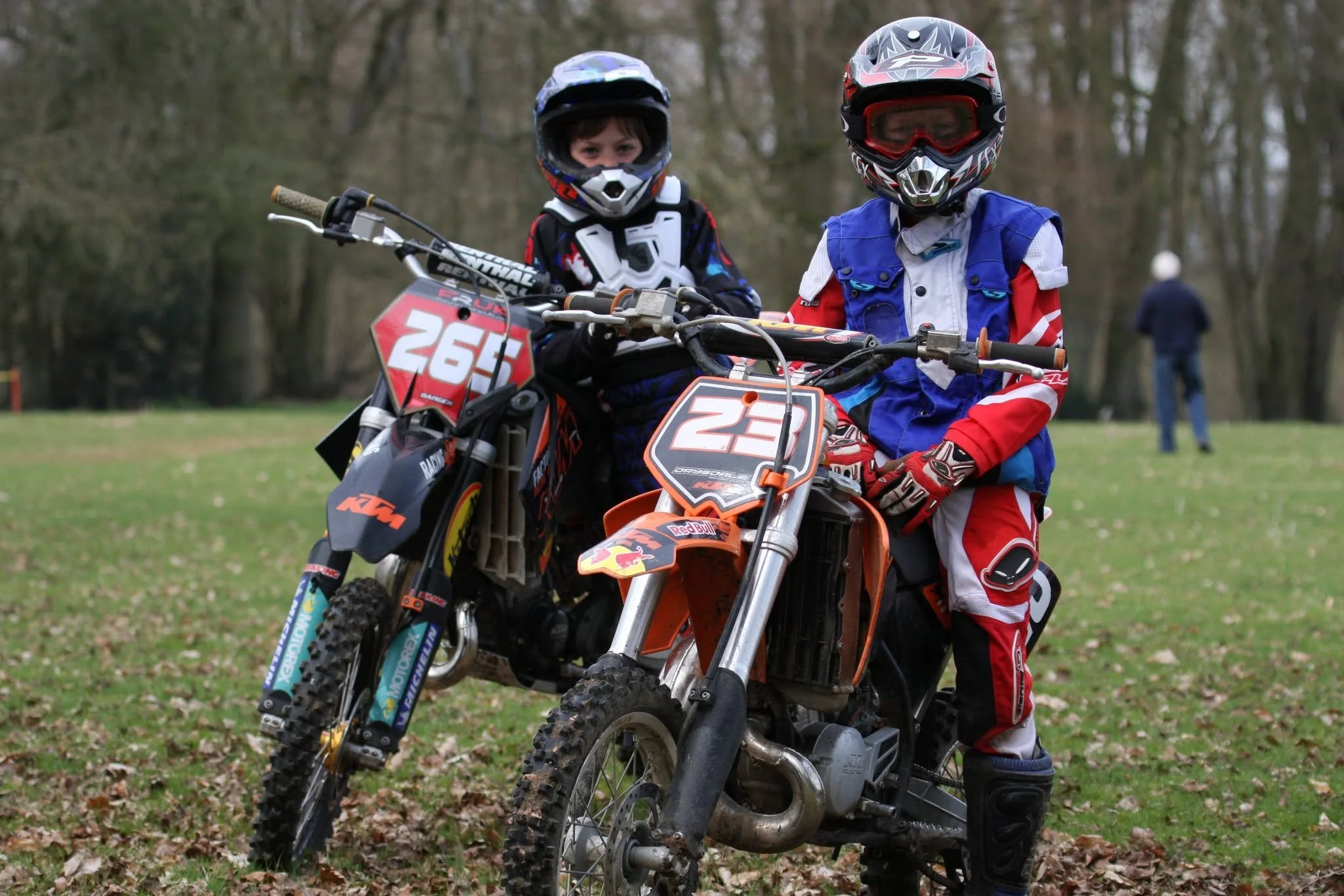 Two children dressed in motocross gear sitting on dirt bikes outdoors, with a person walking in the background.