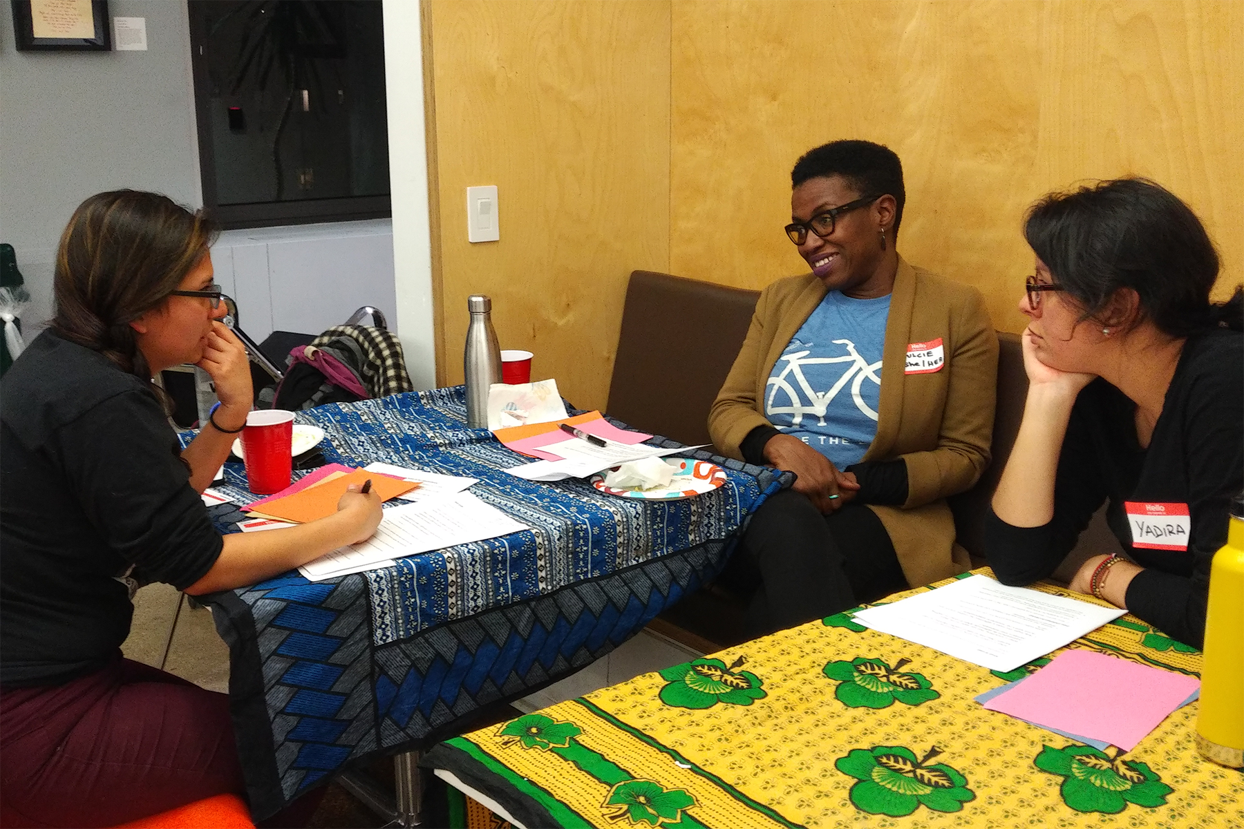 Three women sitting at tables covered with colorful tablecloths in a room, having a discussion or meeting. One woman is taking notes, and another is smiling, wearing glasses and a brown blazer. All three have name tags.