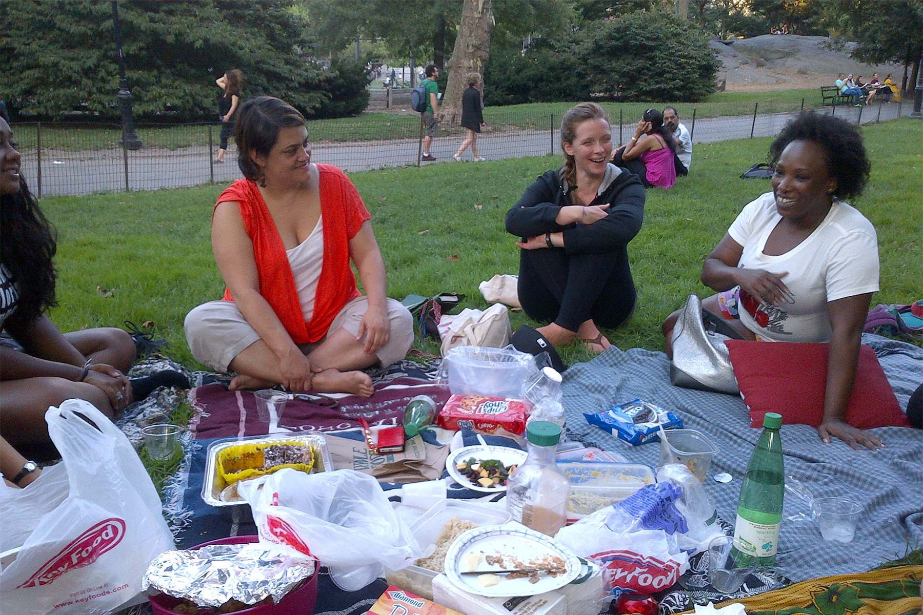 Group of women sitting on blankets having a picnic in a park with food, drinks, and snacks on the blankets, surrounded by trees and other park visitors.