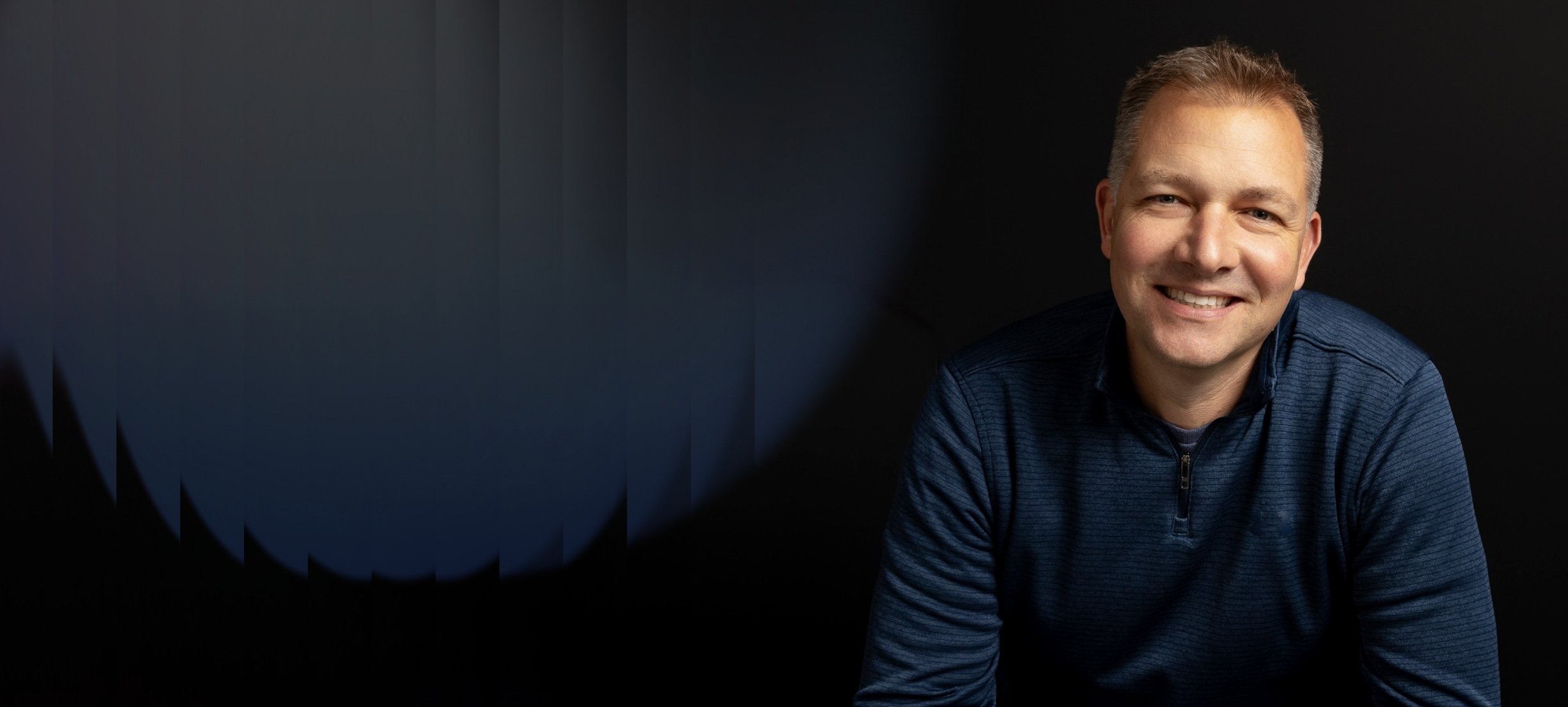 A smiling man with short hair and a dark blue shirt sitting against a dark background.