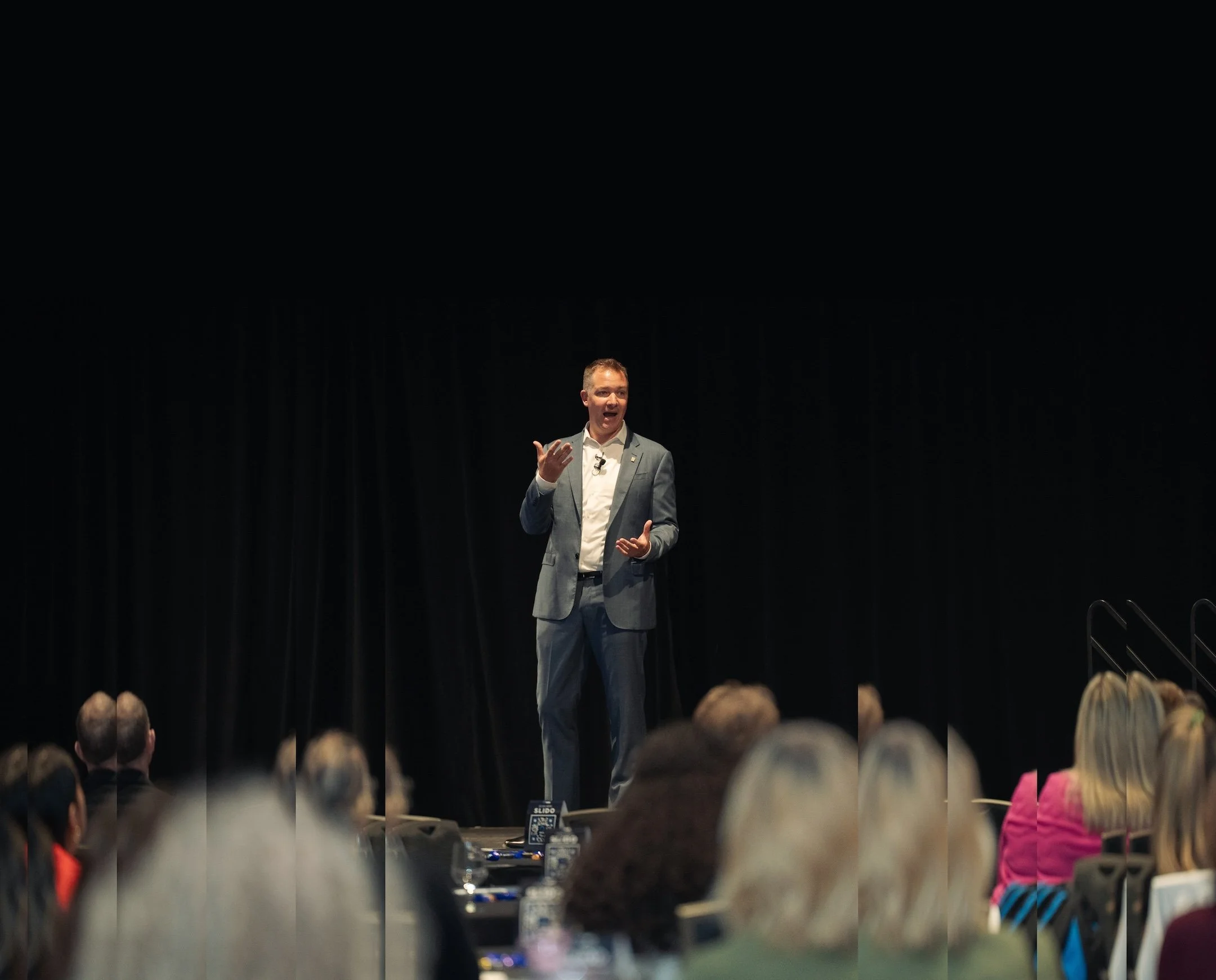 A man in a gray suit is speaking passionately on stage during a presentation or speech, with a dark curtain behind him and an audience watching.