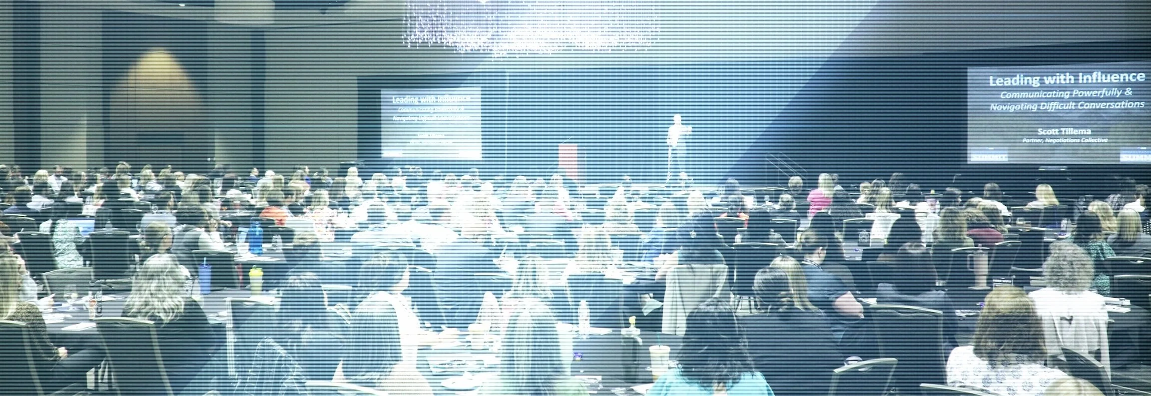 A large conference room filled with seated attendees listening to a speaker on stage. Two screens display the presentation title 'Leading with Influence'.